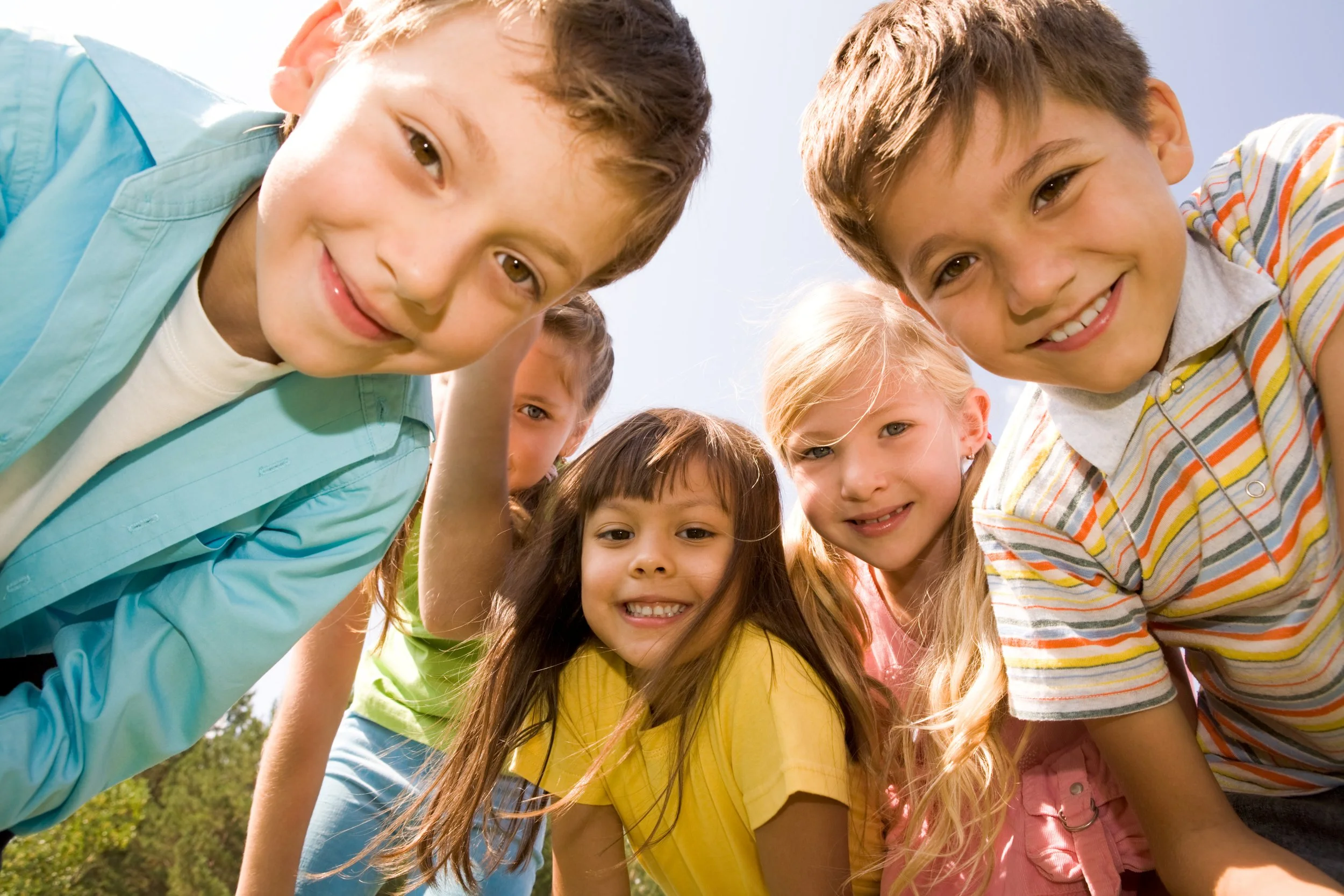 A group of children leaning over, smiling, and looking into the camera during a sunny outdoor day.