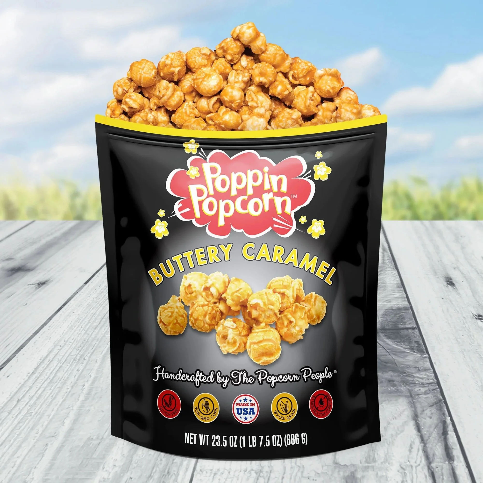 A black bag of Poppi Popcorn Buttered Caramel popcorn with caramel-coated popcorn pieces visible at the top and a bright, outdoor background with blue sky and clouds.