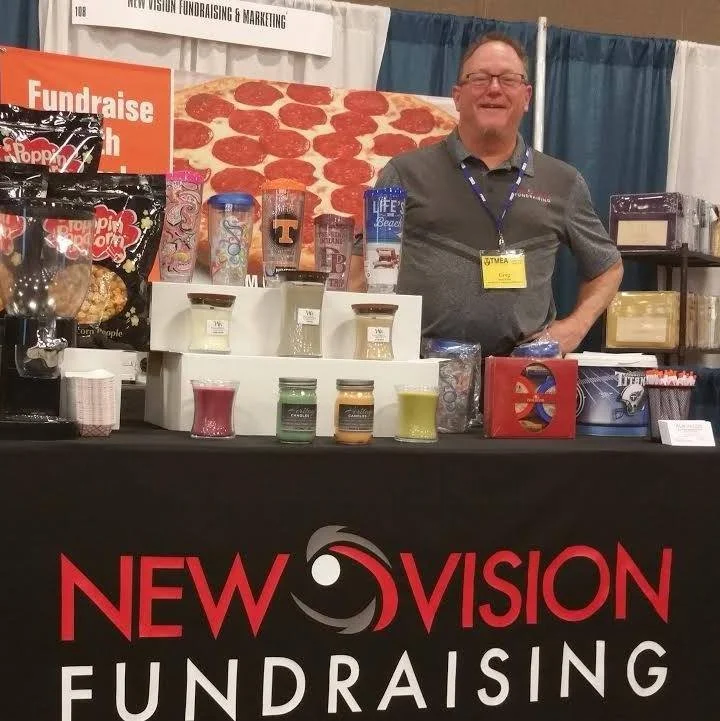 A man standing behind a table with various colorful jar products at a trade show booth for 'New Vision Fundraising.' The booth features a backdrop with pizza imagery and signs for fundraising and marketing.