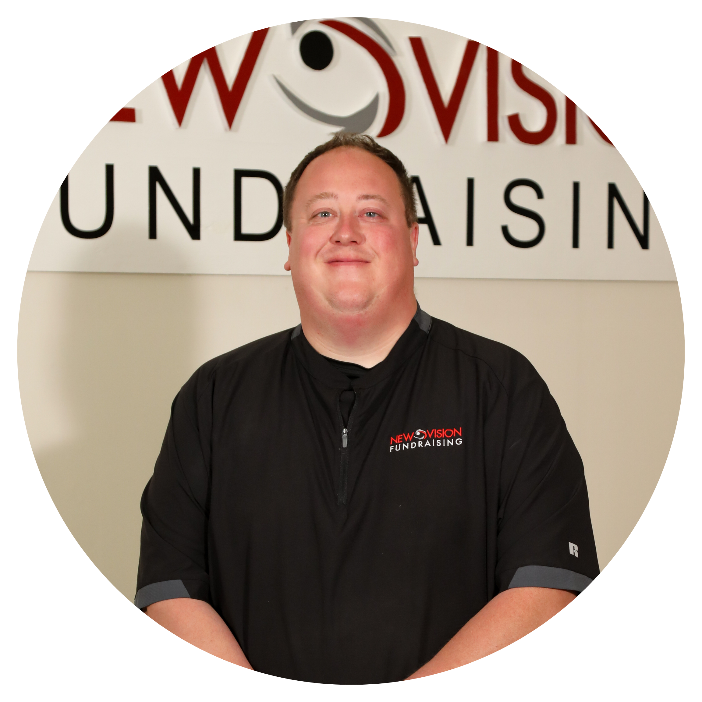 A man with short hair and a wide smile wearing a black shirt with red and white text that reads 'NEW VISION FUNDRAISING' poses in front of a white wall with the partially visible words 'WOR' and 'FUNDRAISING' in black and red letters.