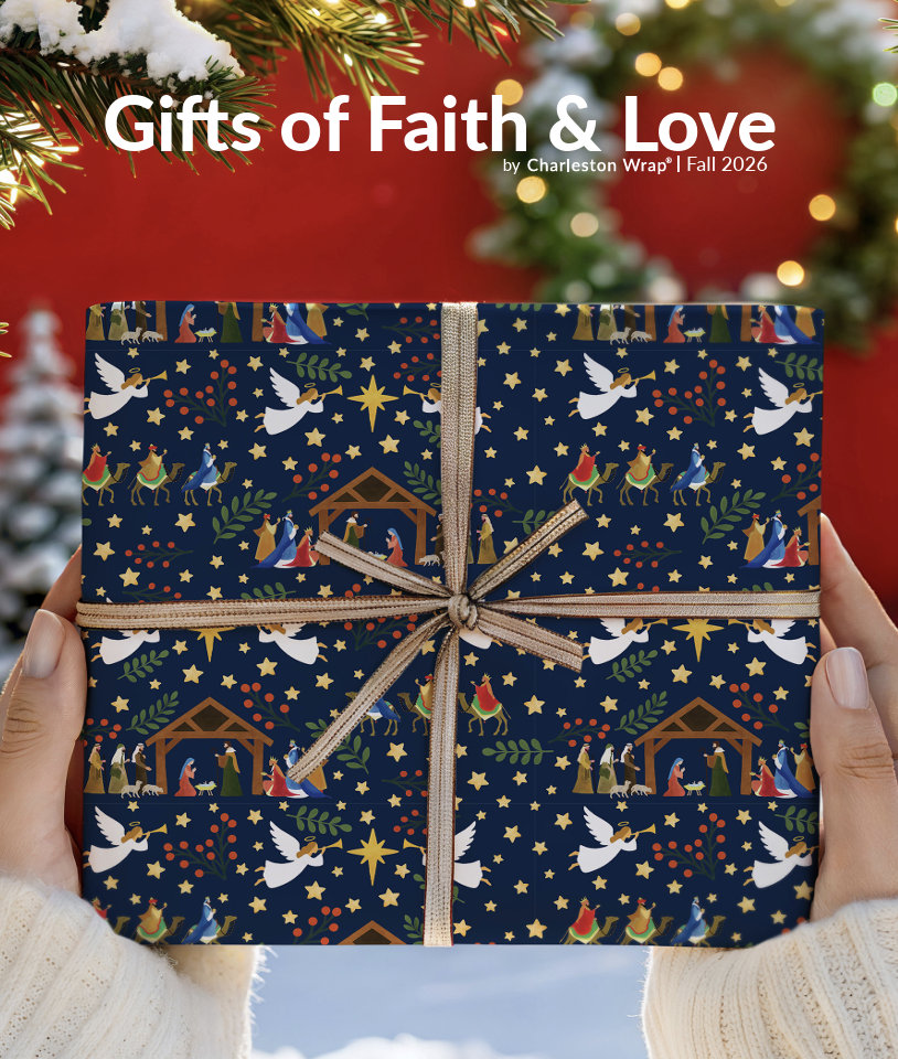 A person holding a Christmas gift wrapped in navy paper with nativity scene, stars, birds, and greenery, tied with a gold bow, in front of a decorated Christmas tree.
