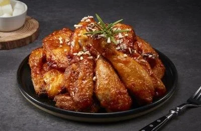 A black plate of crispy fried chicken wings garnished with chopped garlic and a sprig of rosemary, served with a small bowl of dipping sauce in the background.