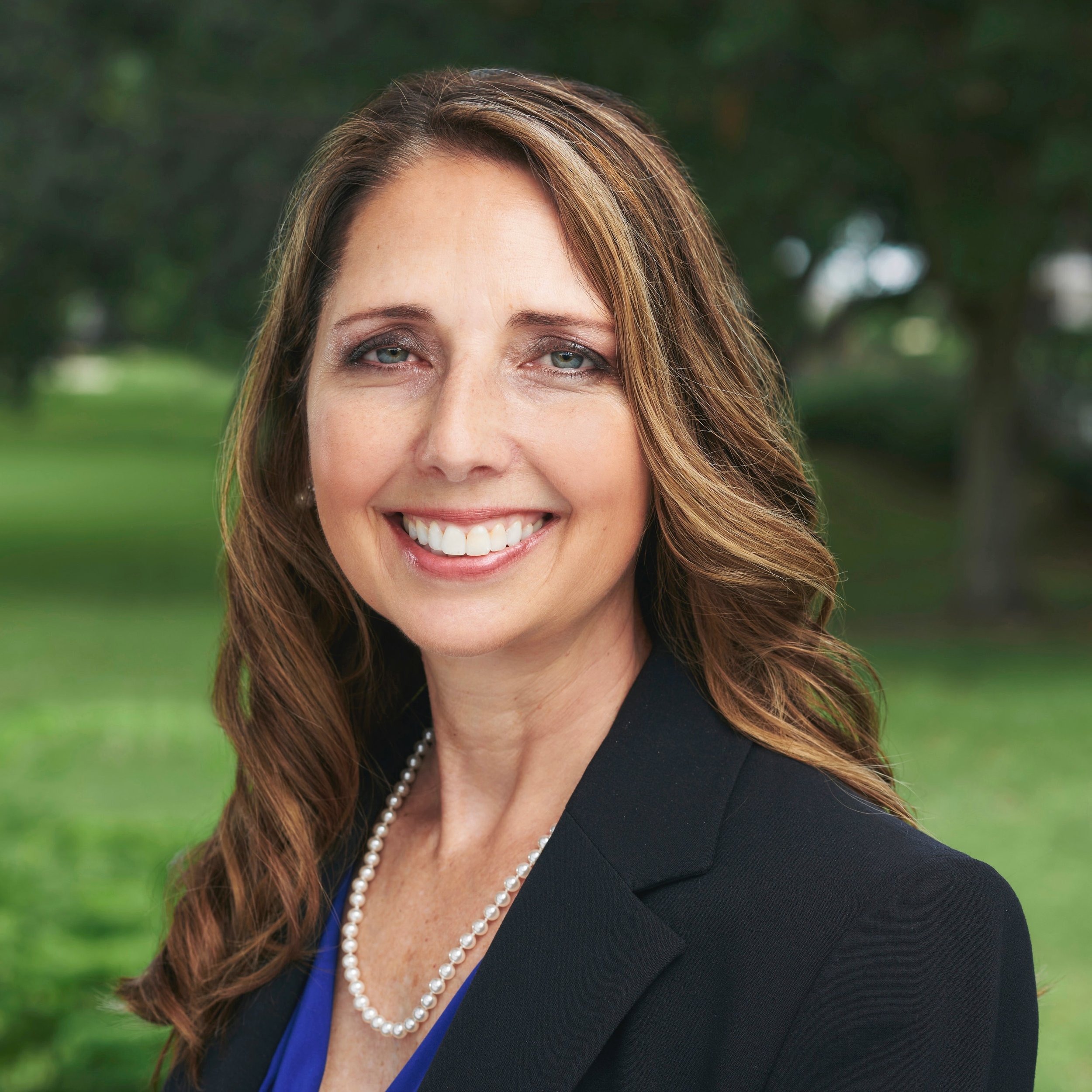 Portrait of a smiling woman with wavy brown hair, wearing a black blazer and pearl necklace, outdoors with blurred green trees in the background.