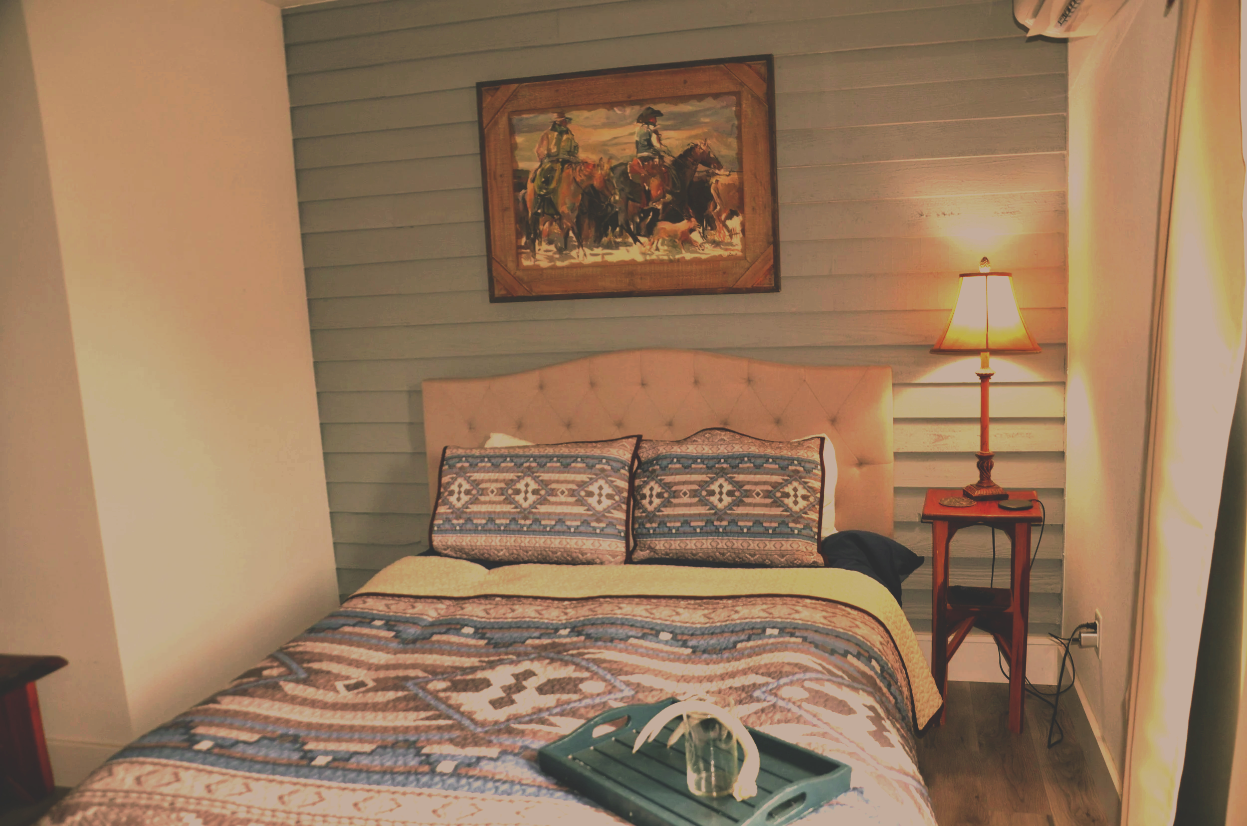 The bedroom features a bed with a patterned quilt and matching pillows, a wooden nightstand with a table lamp, a framed picture of cowboys riding horses hanging on the wooden-paneled wall, and a window with curtains on the right side.