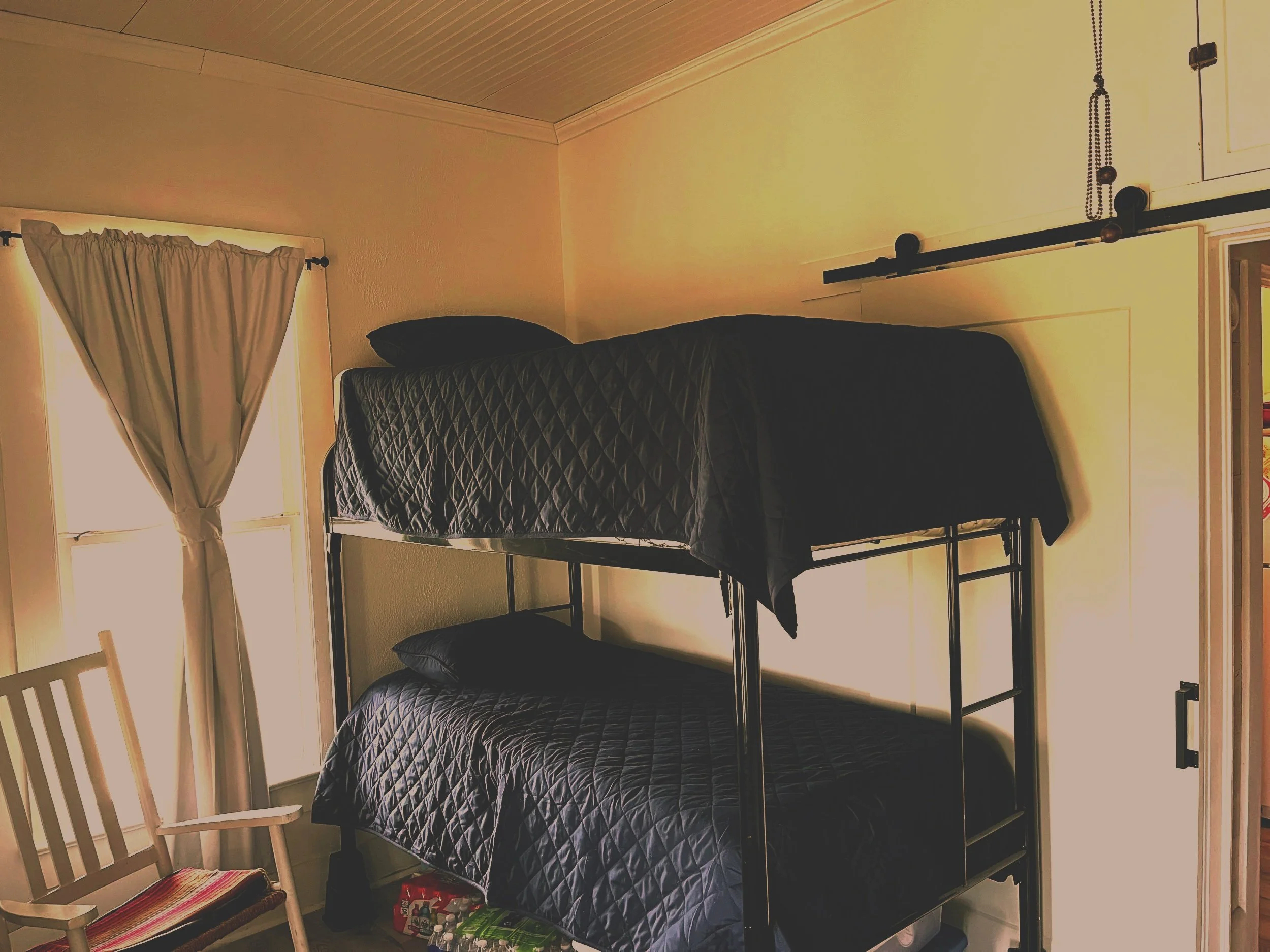 A room with metal bunk beds with black quilted bedding, a wooden chair with a colorful cushion, and a window with beige curtains. It has light-colored walls and ceiling with wood paneling.