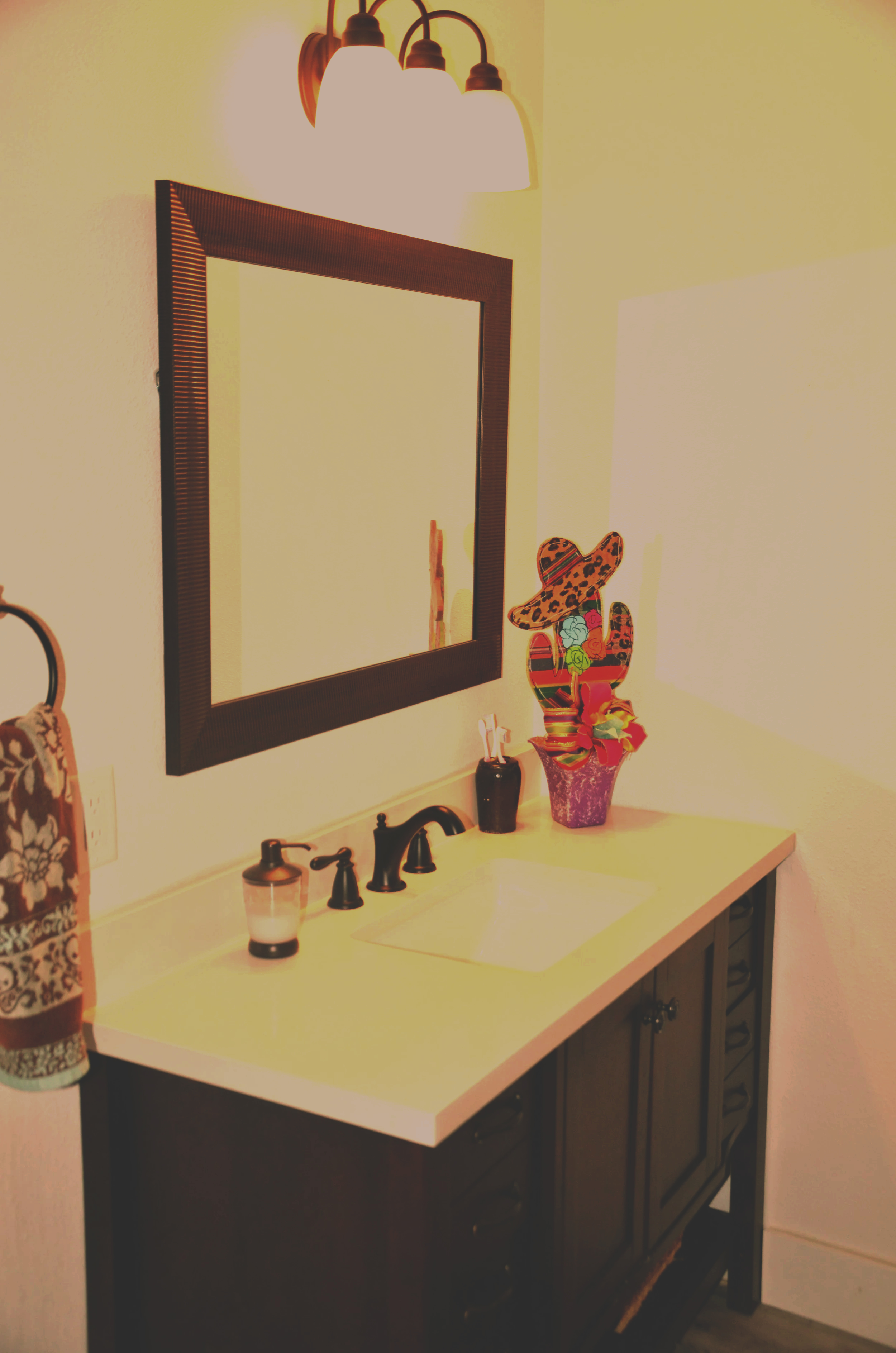A bathroom vanity with a dark wooden cabinet and white countertop, topped with a decorative cactus in a pink pot with a large sombrero and colorful ribbon. Above the vanity is a mirror with a wooden frame, and to the left are some wall decorations and a soap dispenser.