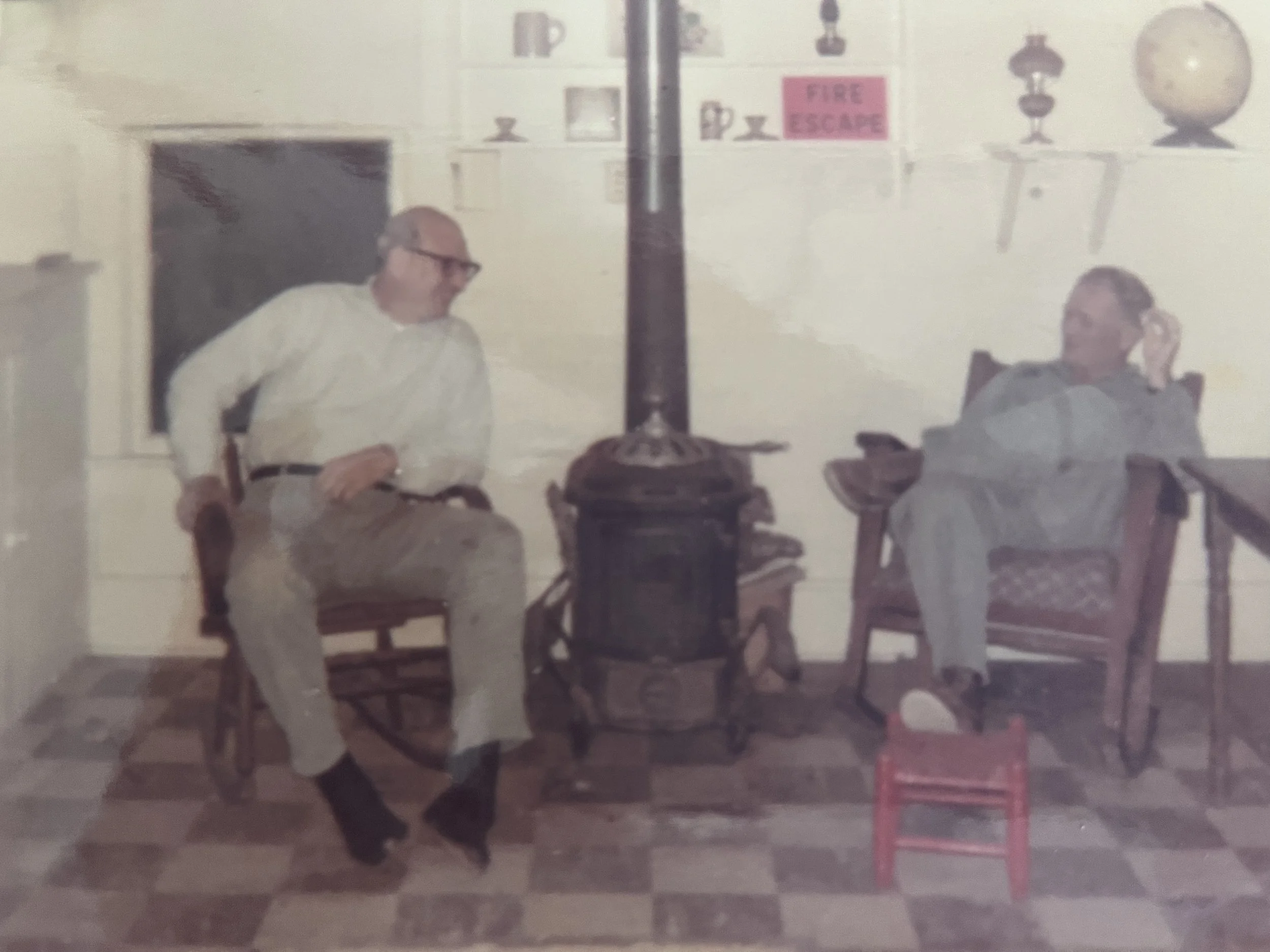Two men are sitting and talking in a room. The man on the left is wearing glasses and a light-colored shirt, while the man on the right is wearing a darker shirt. A small table is placed between them, with a lamp on it. Behind the men, there is a wall with some shelves, decorative items, and a pink sign that reads 'FIRE ESCAPE'. There's a red chair or display stand on the right side of the image.