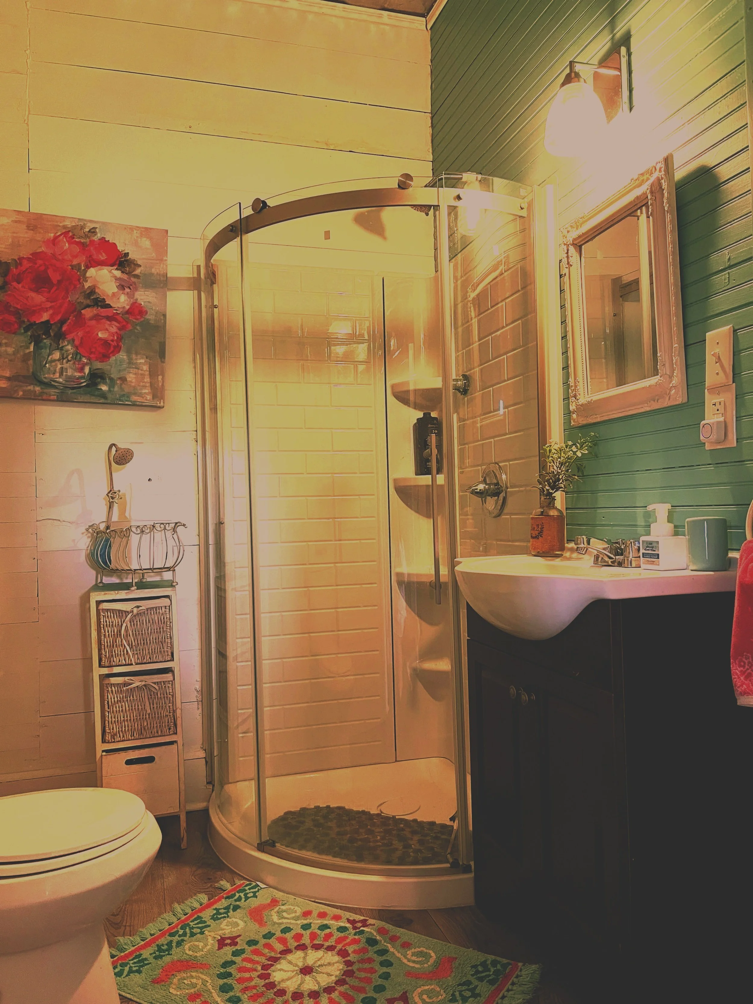 A bathroom with a corner shower, a toilet, a small cabinet, a sink with a mirror, and various decorations including a painting of red flowers and a colorful rug.