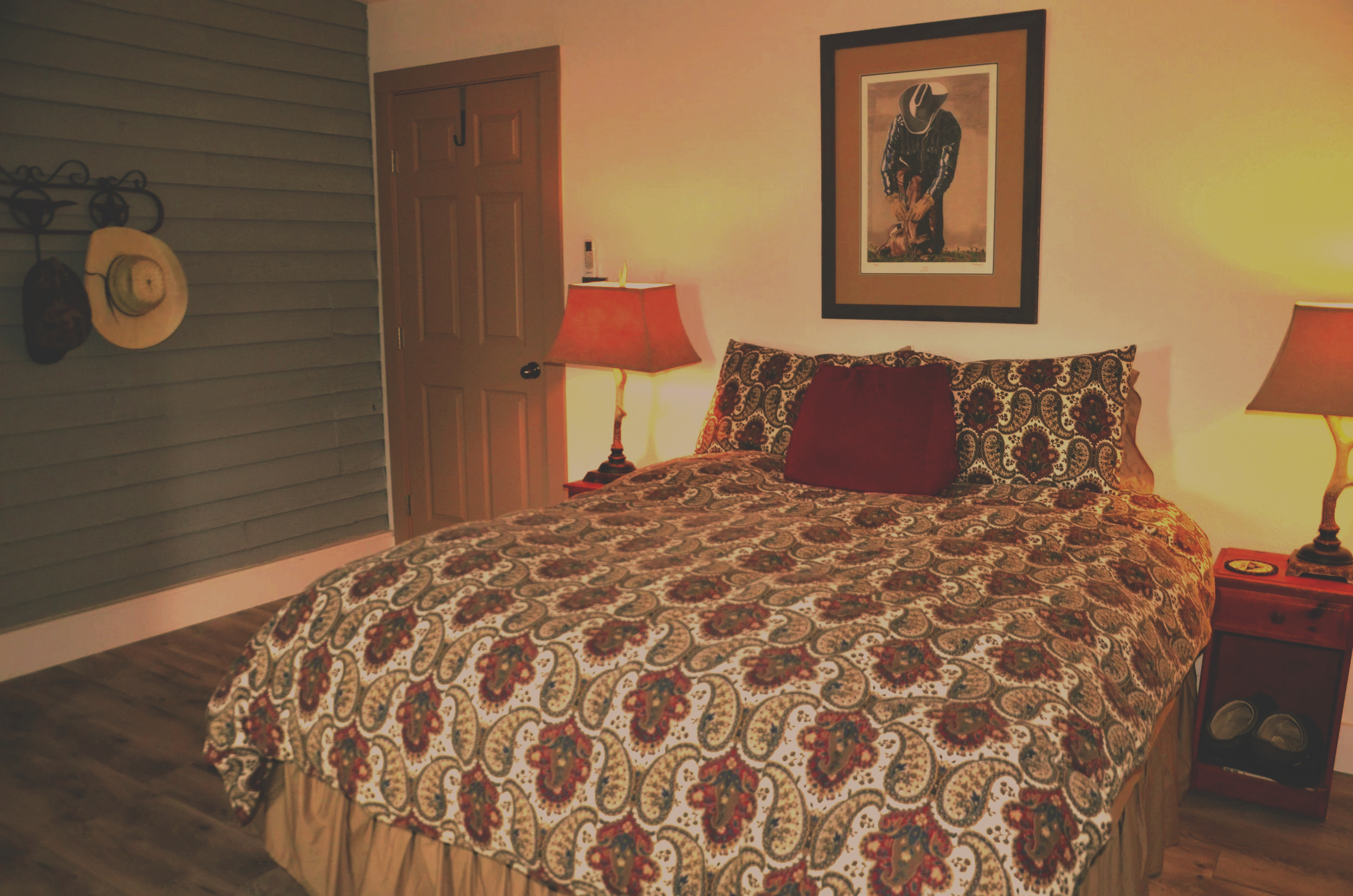 A bedroom with a bed covered in a patterned quilt and pillows, two nightstands with lamps, a framed cowboy picture on the wall, a wooden door, and a wall with hats hanging on hooks.