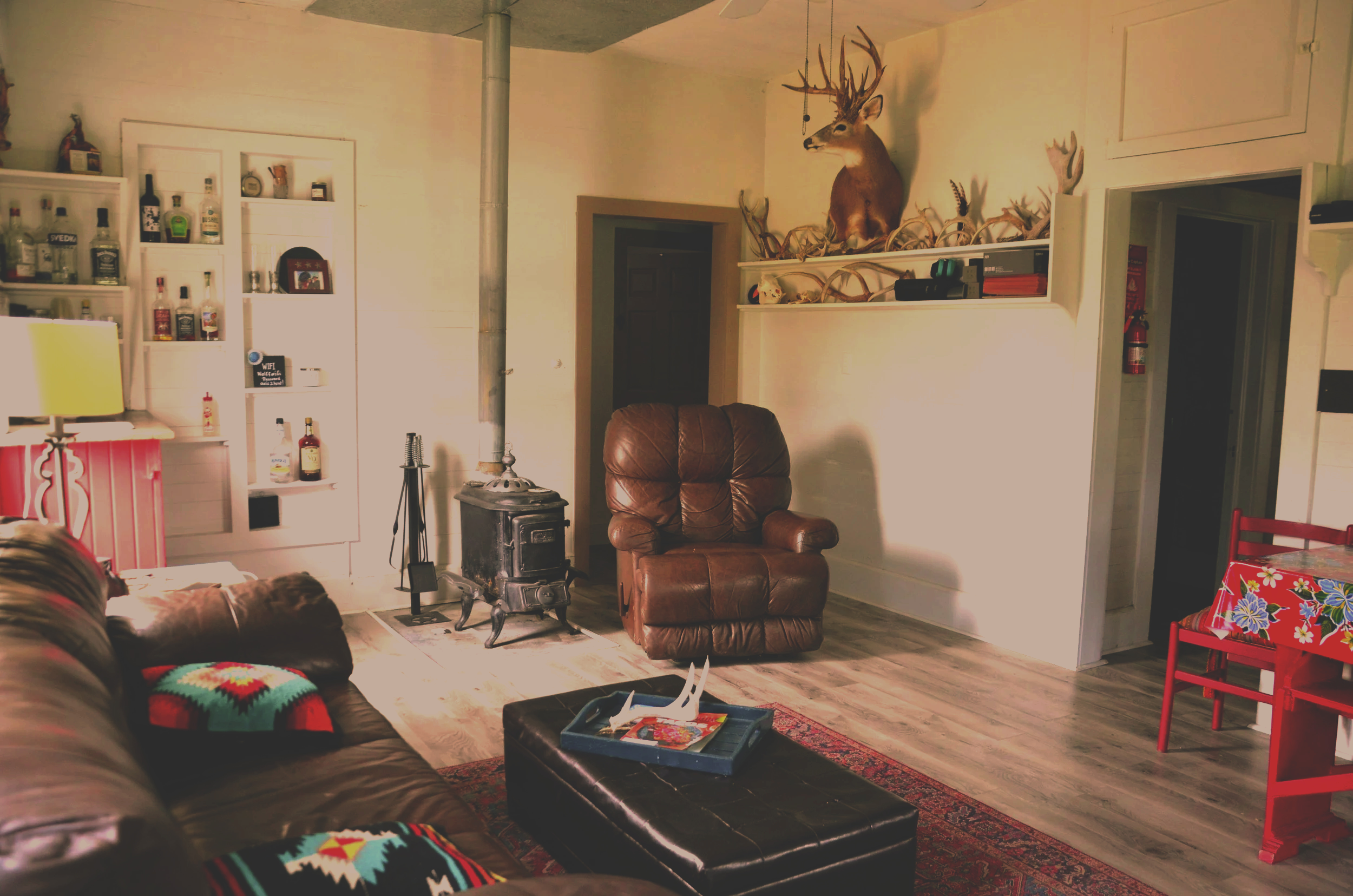 Living room with a leather sofa, a small black wood stove, a brown leather armchair, a red chair, a white shelving unit with alcohol bottles, a colorful crocheted blanket on the sofa, and a deer head mounted on the wall above a shelf decorated with antlers.