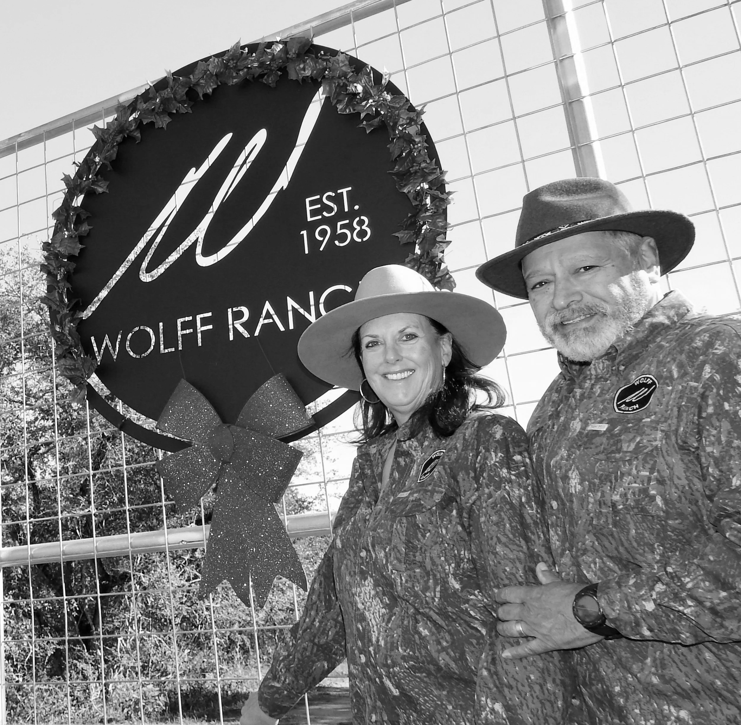 Black and white photo of a person with glasses and a hat, smiling, standing in front of a sign that reads 'EST. 1958 WOLFF RANCH' decorated with leaves and glitter, with a glass building in the background.