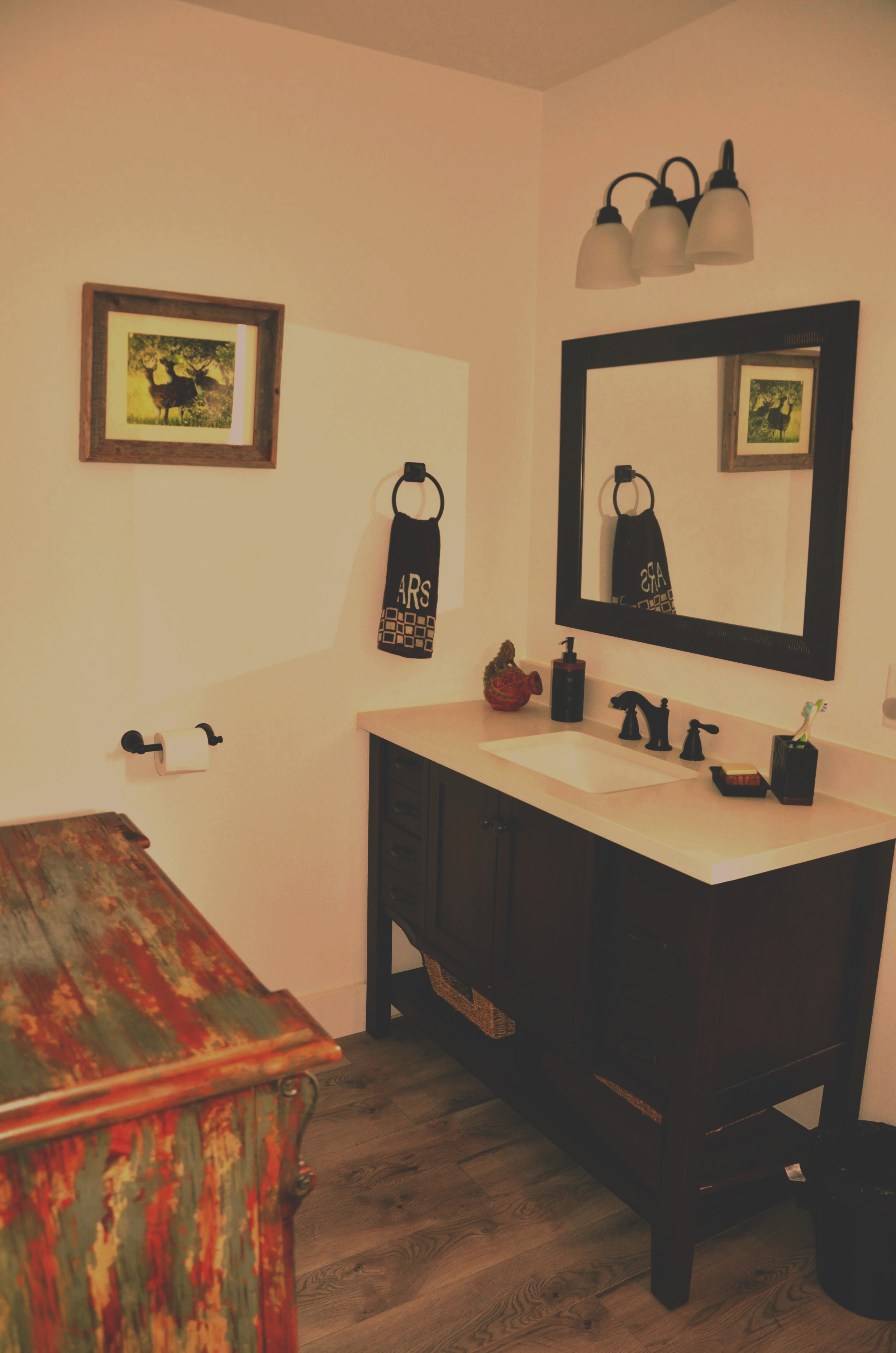 A bathroom vanity with a white countertop, black fixtures, and a large black mirror. A framed picture of deer in a field hangs on the wall. A black towel with white lettering is on a towel ring, and a small decorative object and a soap dispenser are on the counter. Part of a colorful wooden table is visible in the foreground.