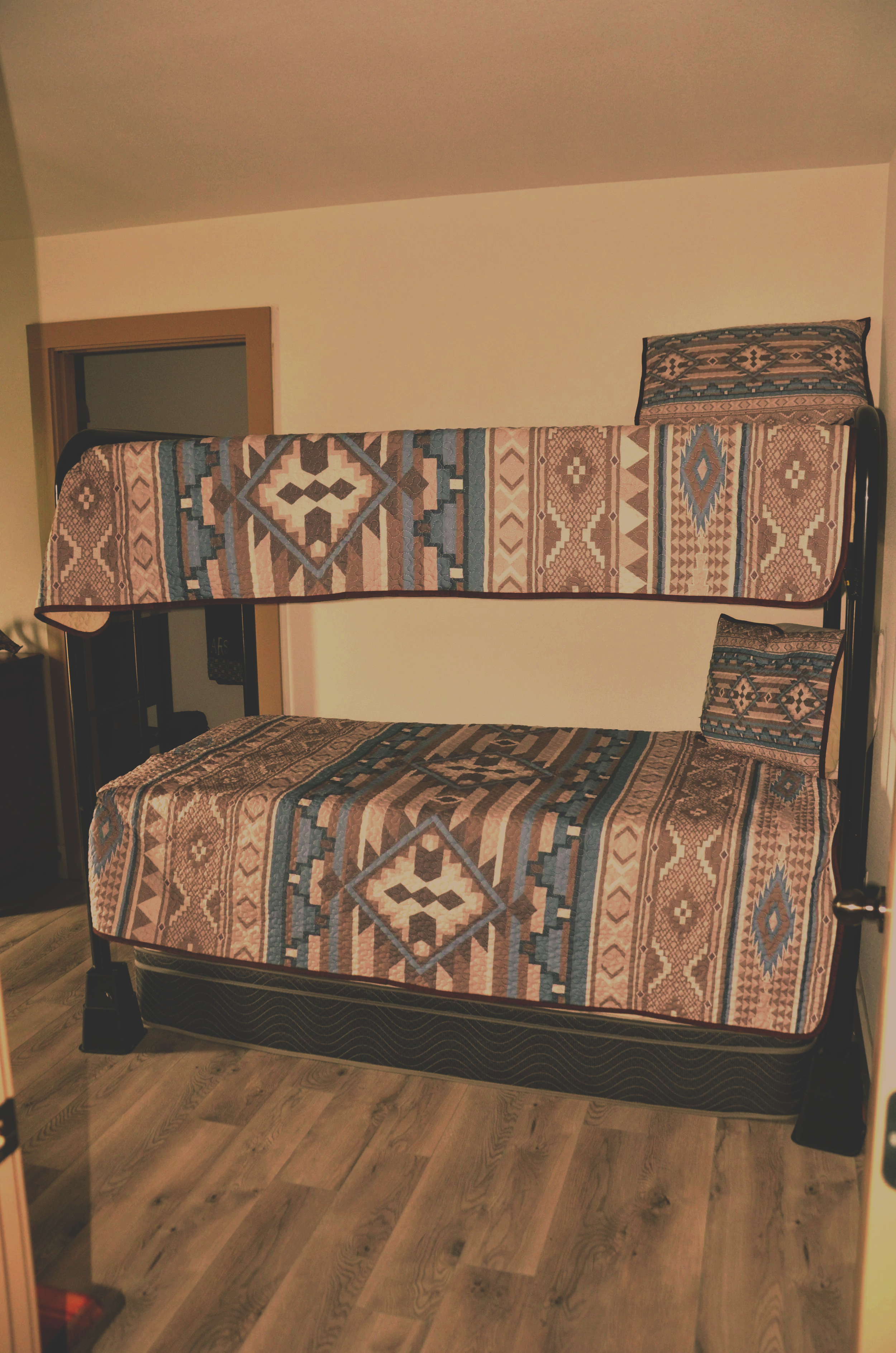 A bunk bed with patterned bedspreads and pillows, situated against a beige wall in a room with wooden flooring.