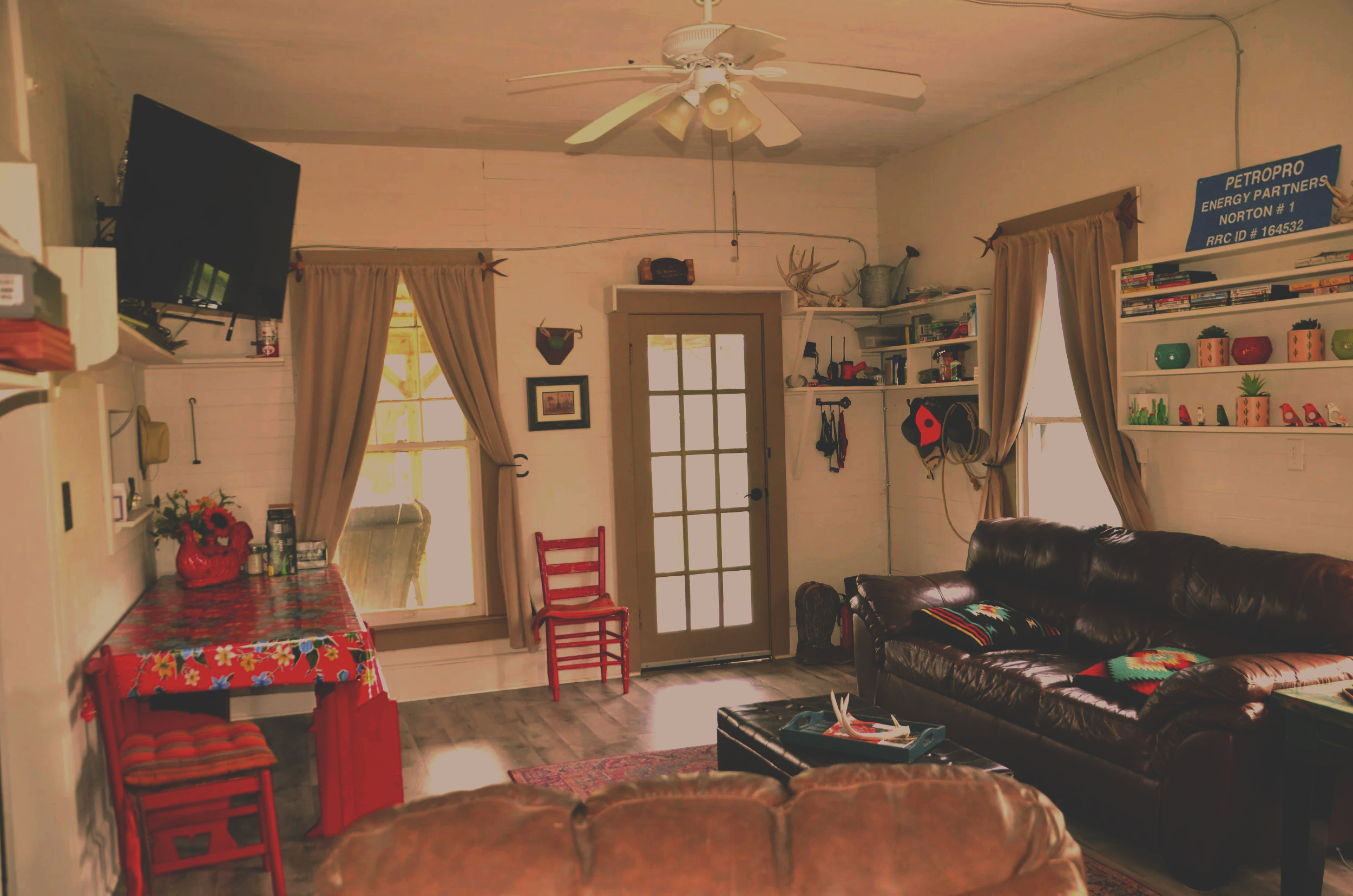 Interior of a cozy living room with white wooden walls, a ceiling fan, a wall-mounted TV, and a door with glass panels. There is a dark leather couch, a red chair, a small table with a tray, and shelves with books and decorative items. Two windows with beige curtains let in natural light.