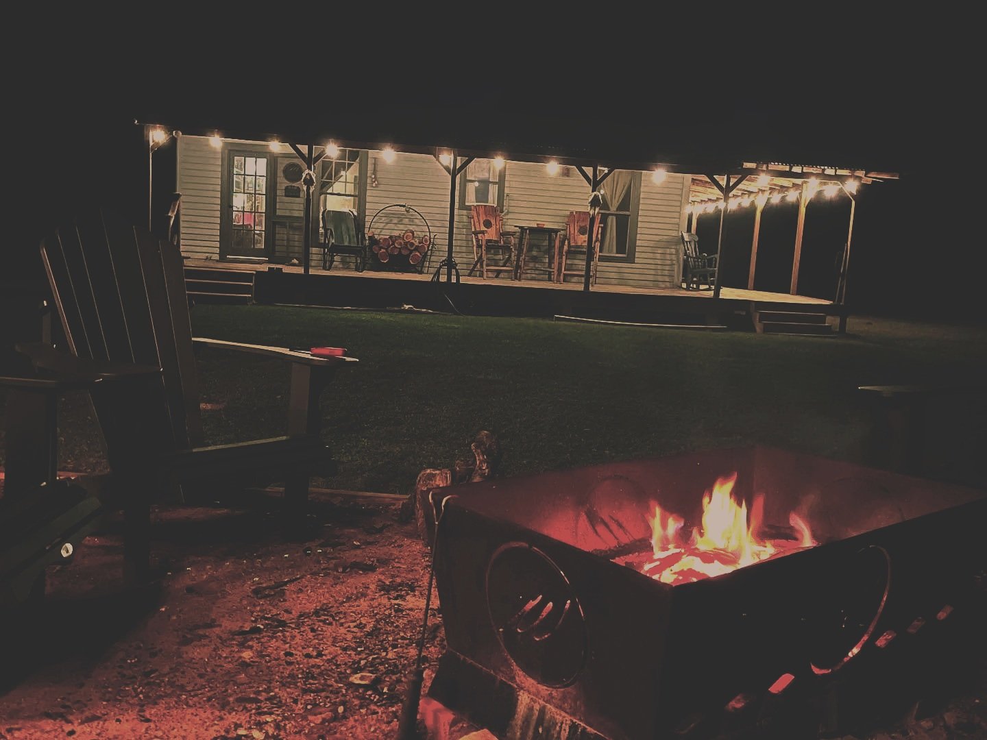Nighttime outdoor scene with a lit fire pit, Adirondack chairs, and a house with a wooden porch decorated with string lights.