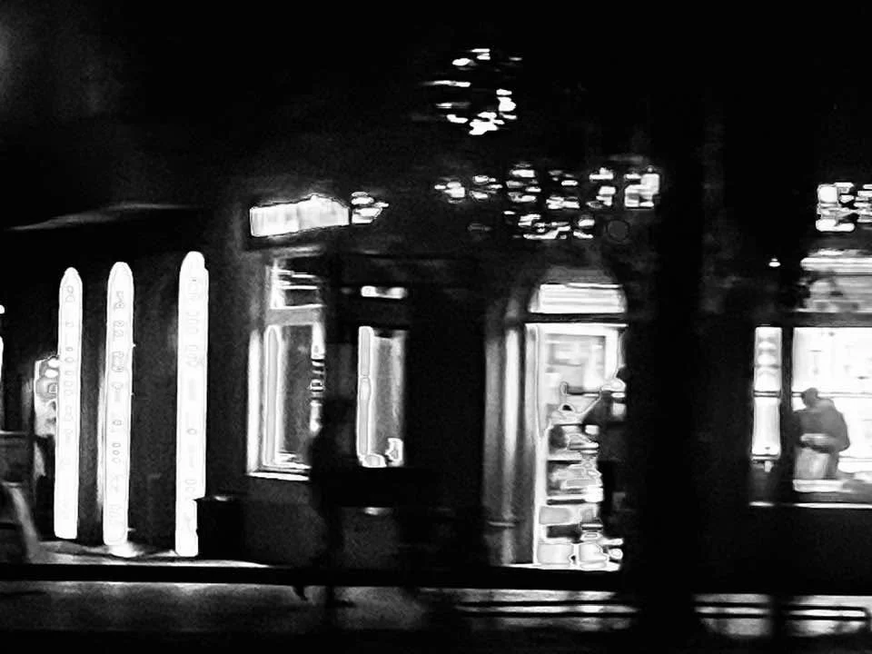 Schwarz-weiß-Fotografie eines beleuchteten Cafés bei Nacht mit Personen im Inneren, die an den Fenstern sitzen.