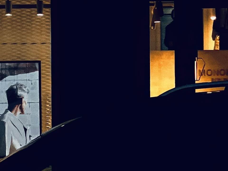 Ein Mann in weißem Kleid und Kochmütze steht vor einem Fenster, im Hintergrund verschwommene Personen im Schatten.