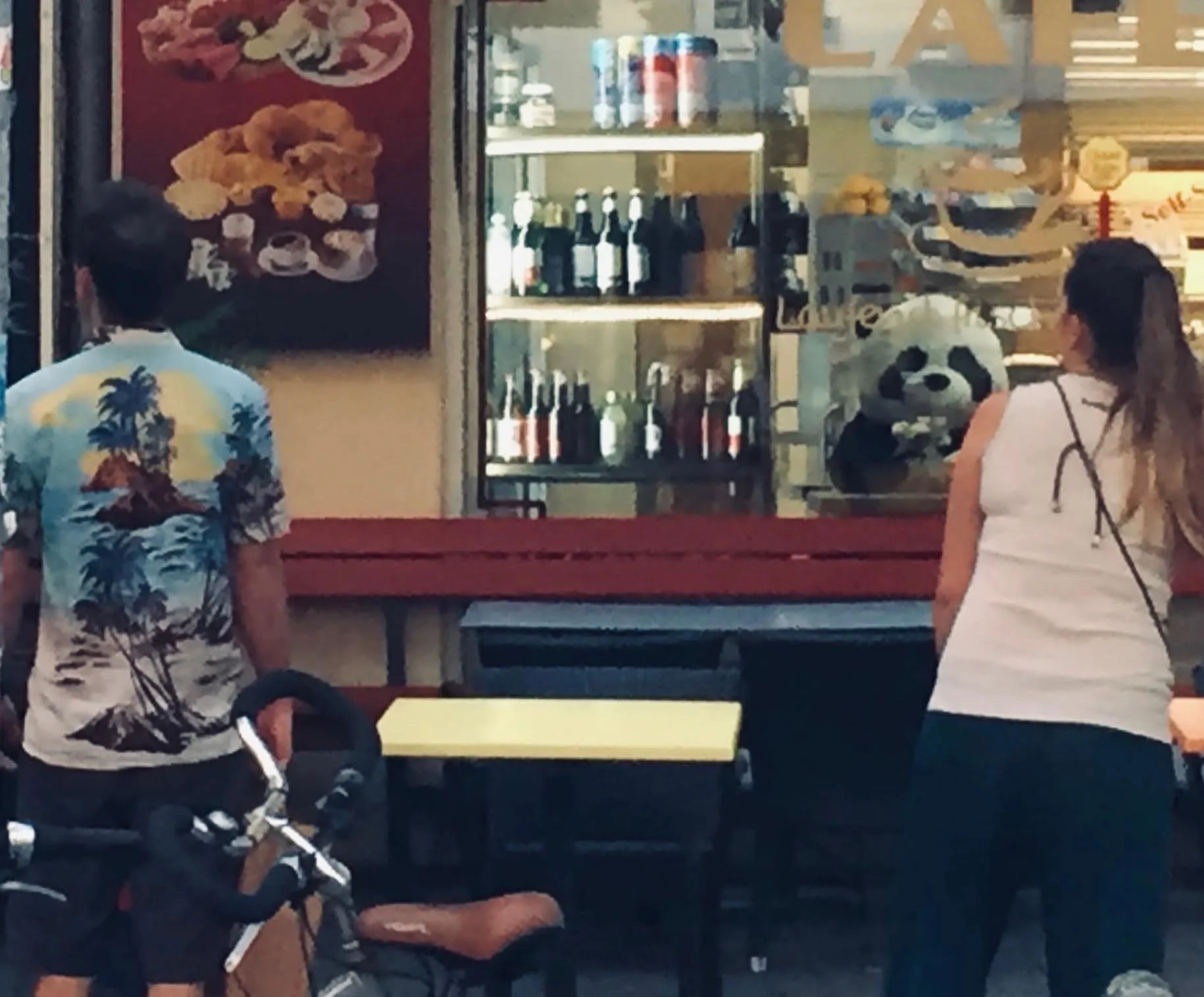 Sich im Inneren eines Restaurants befindende Personen, darunter eine Frau mit einem weißen Top, ein Mann mit einem anderen Top mit einem Palmen-Motiv, eine Frau mit einem weißen Top, die einen Kinderwagen schiebt, und eine Person auf der linken Seite
