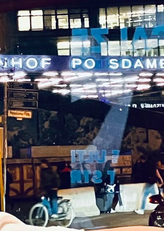 Reflexion eines leuchtenden Schildes mit einem halbleserlichen Text, Menschen, Fahrrad und eine Tasche an einem Busbahnhof bei Nacht