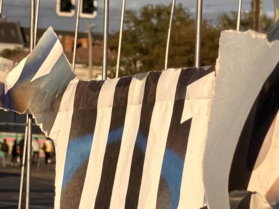 Zerrissene Plakate hängen an einem Metallzaun, im Hintergrund sind Menschen und Gebäude sichtbar.