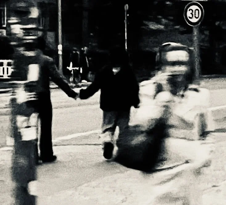 Zwei Männer halten sich an einer Straße nachts an den Händen, eine Person läuft vorbei auf einem Trolley, im Hintergrund Verkehrsschilder und Straßenlaternen.