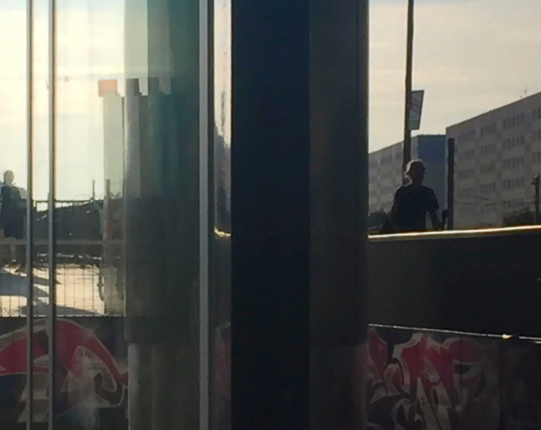 Blick durch eine Glasfassade auf zwei Personen, die auf einer Straße stehen, und auf ein städtisches Gebäude im Hintergrund, bei Sonnenuntergang oder Sonnenaufgang.