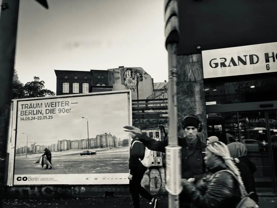 Schwarz-weiß-Foto einer Berliner Straße mit Passanten, einer großen Plakatwand und teilweise sichtbareren Gebäuden im Hintergrund.