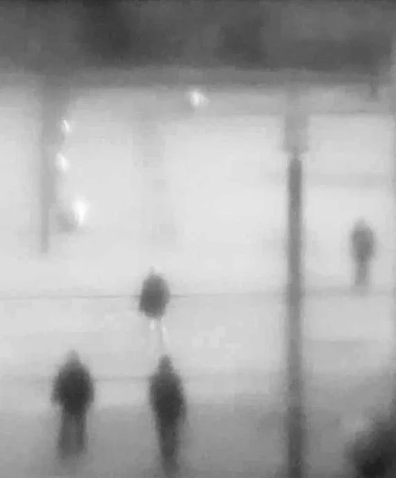 Schwarz-weiß-Foto einer Person auf regennasser Straße bei Nacht, beleuchtet von Straßenlaternen.