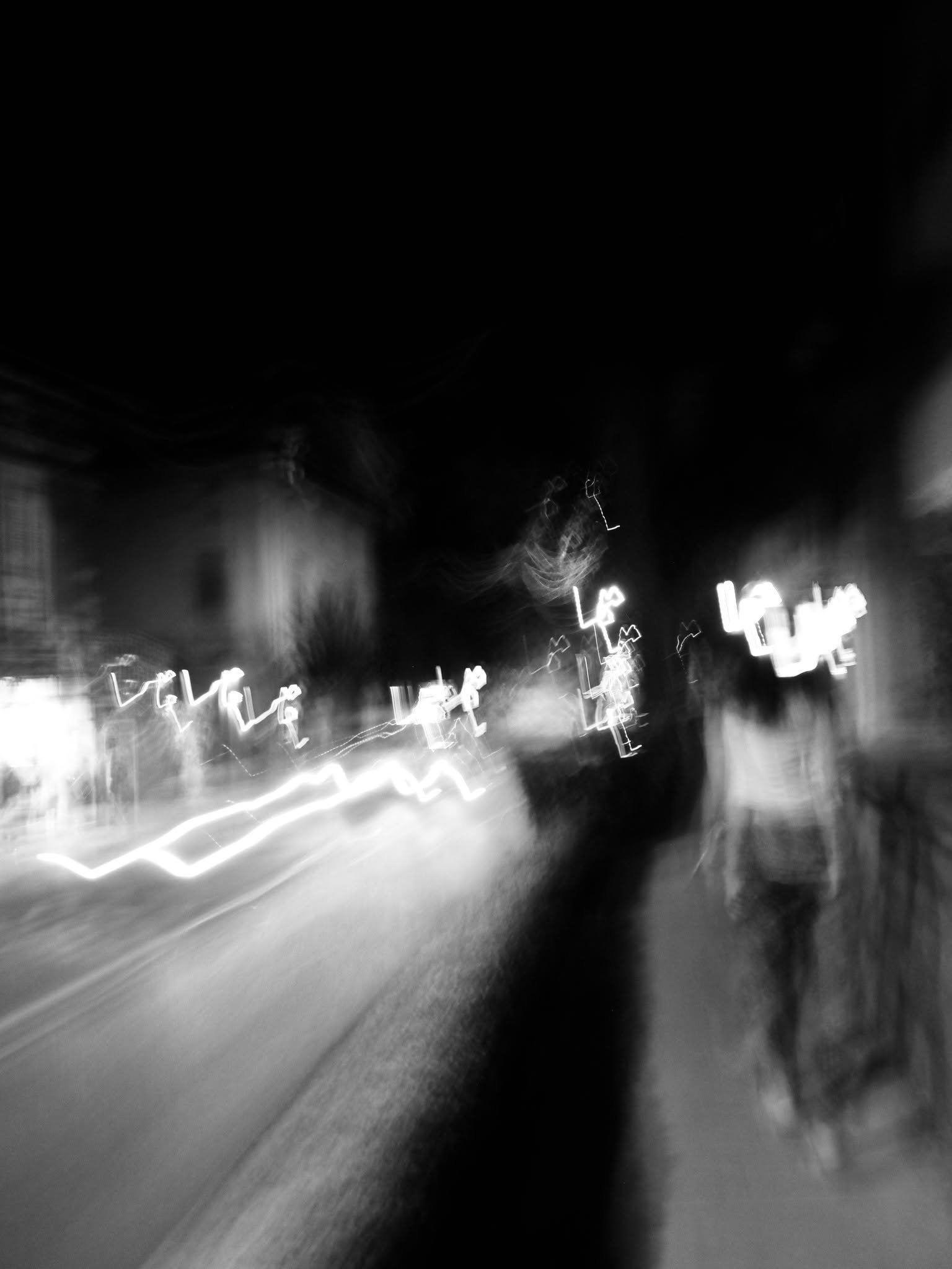Unscharfes Schwarz-Weiß-Foto einer nächtlichen Straße mit verschwommenen Lichtern und Gebäuden.