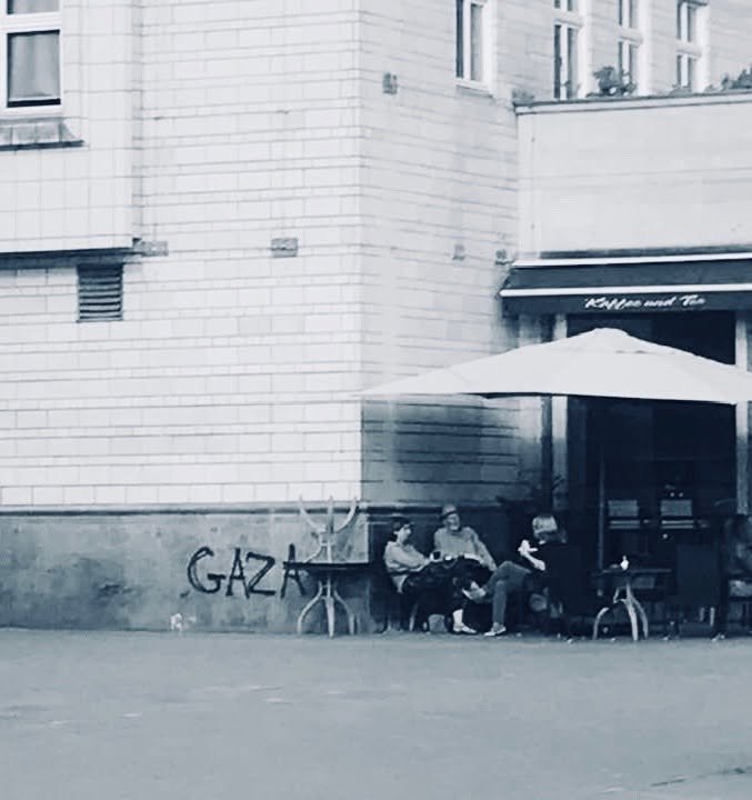 Drei Menschen sitzen an einem Tisch vor einem Café mit großem Schirm unter einem Graffiti am Gebäude.