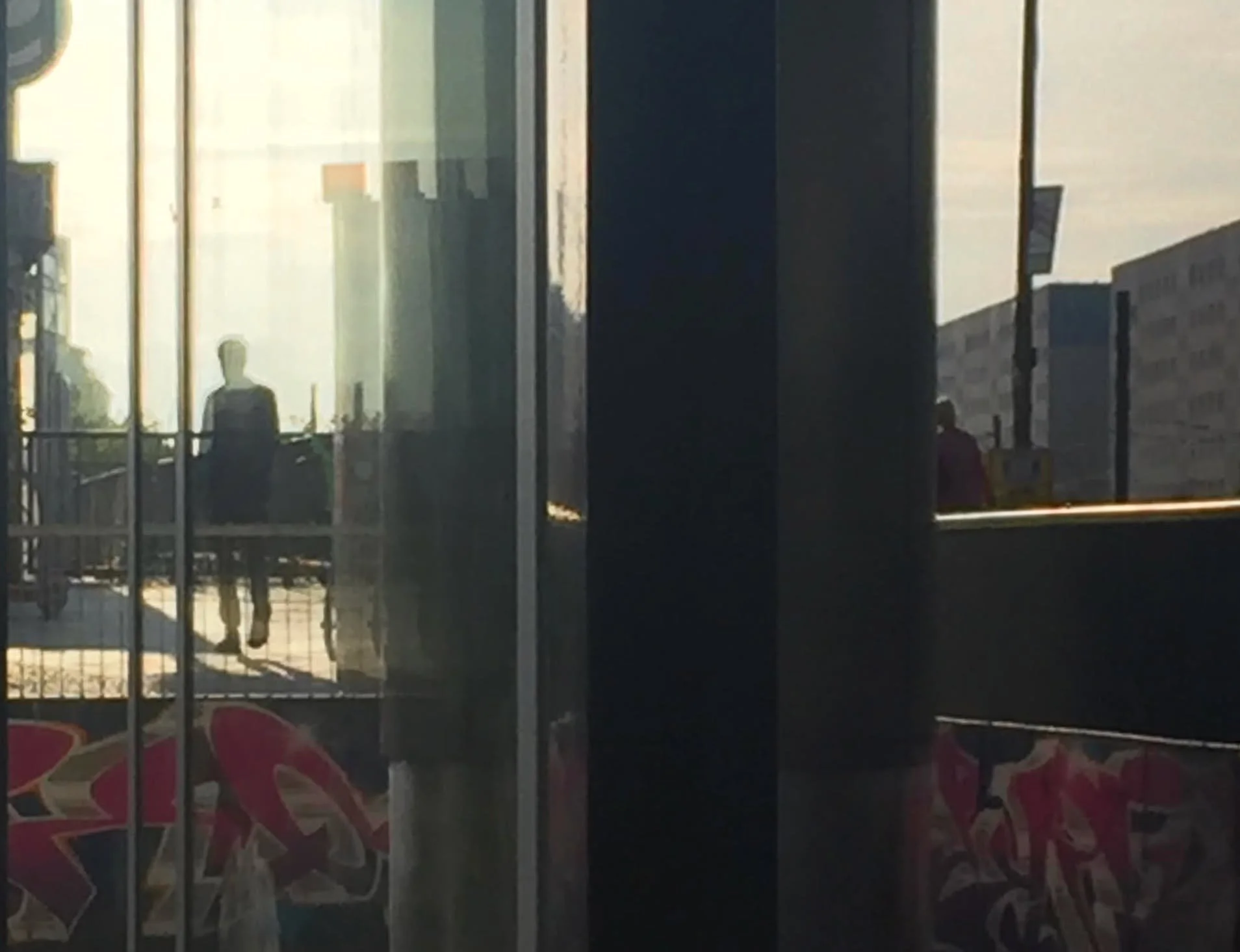 Reflexion eines Mannes, der mit Maske unterwegs ist, durch eine Glasfassade, mit Graffiti auf der unteren Ebene, im Hintergrund Gebäude und Sonnenschein.