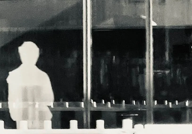 Ein verschwommenes Bild mit einer weißen Silhouette einer Person vor dunklem Hintergrund, möglicherweise in einem Geschäft oder Lager mit Regalen.