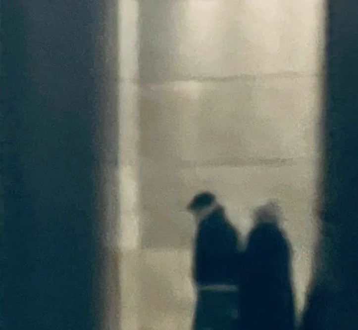 Das Bild zeigt zwei Menschen, die in einem Flur oder Korridor nebeneinander gehen, mit einer Wand im Hintergrund.