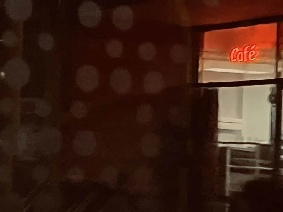 Ein dunkler Raum mit einer roten, leuchtenden Café-Schrift an der Wand und einem Fenster, durch das eine helle Inneneinrichtung sichtbar ist.