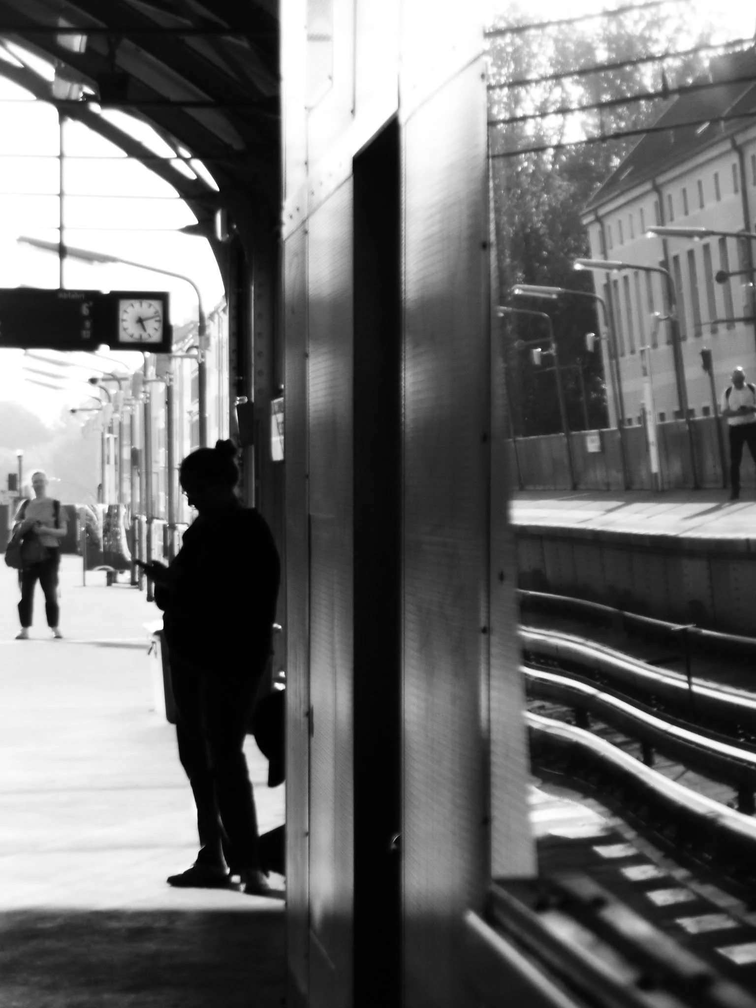 Menschen warten an einem Bahnsteig, einige lesen oder schauen auf ihre Handys, ein Bahnhof mit Uhren ist sichtbar.
