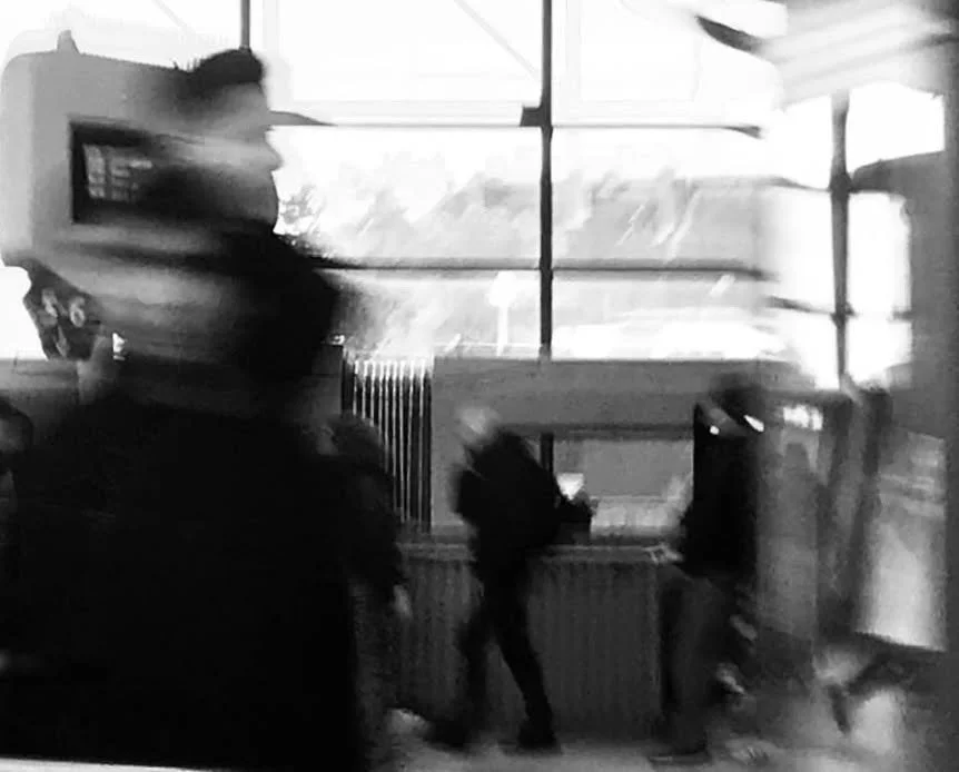 Schwarz-weiß-Fotografie in einem öffentlichen Raum, mit mehreren Personen, die vor einem Fenster stehen, das den Blick nach draußen freigibt.