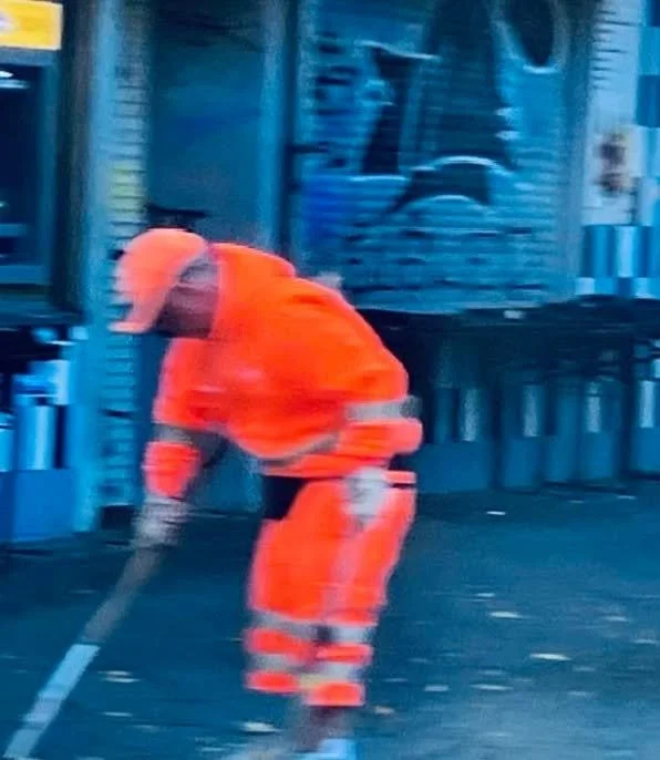 Straßenarbeiter in orangefarbener Warnbekleidung arbeitet auf der Straße, vermutlich bei Straßenreparatur oder -reinigung.