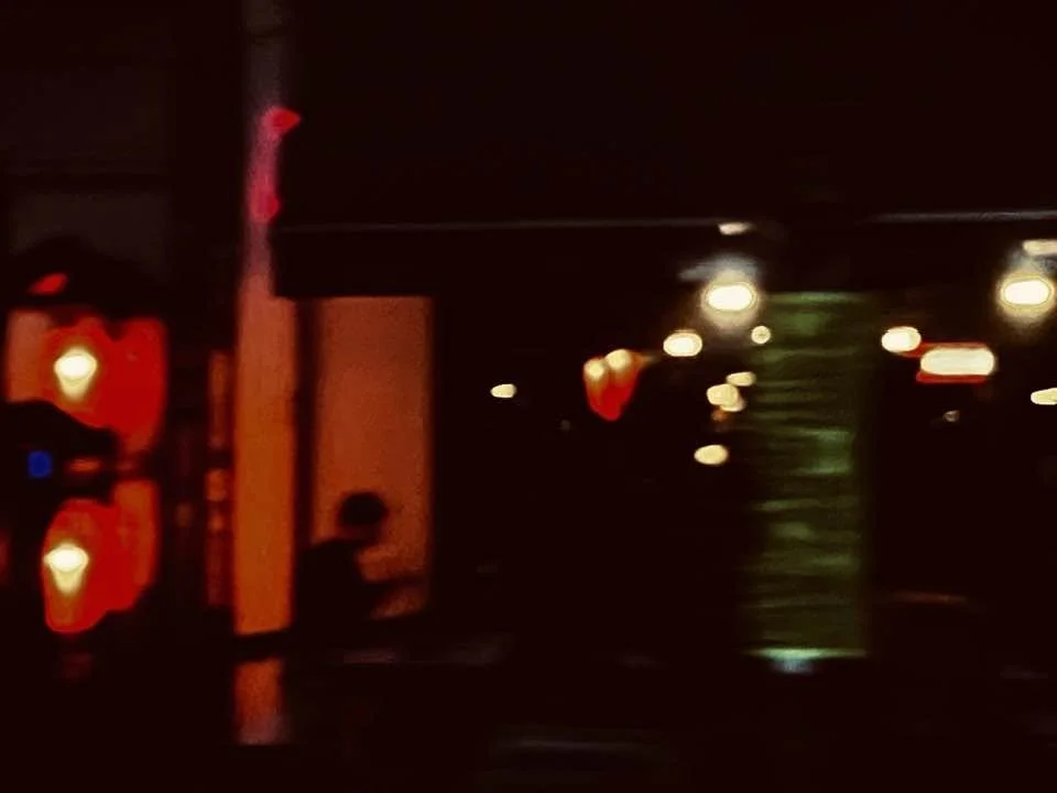 Ein unscharfes, dunkles Bild mit verstreuten Lichtern und einer Silhouette im Vordergrund. Es sieht nach einer nächtlichen Szene aus, möglicherweise in einer Stadt oder auf einer Straße.