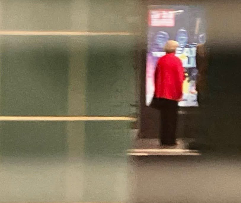 Eine Person in rotem Mantel steht vor einem Getränkeautomaten in einem dunklen Raum oder Flur.
