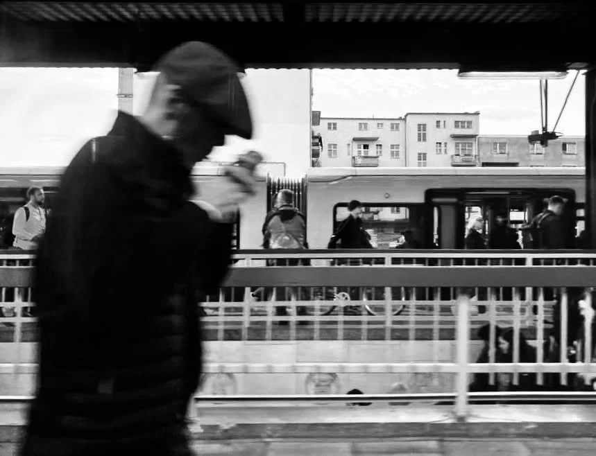 Schwarzweißbild von einer Person, die einen Regenschirm hält und vorbeigeht, während im Hintergrund Menschen an einer Haltestelle warten und ein Bus steht.
