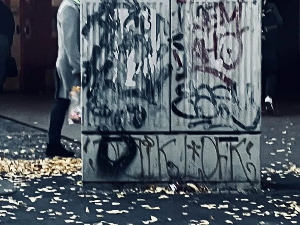 Graffiti an einer Wand, Menschen im Hintergrund, Blätter auf dem Boden.