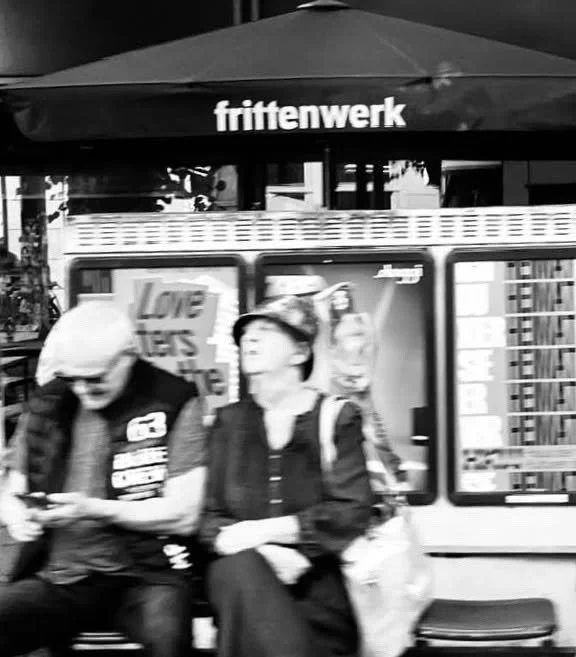 Zwei Menschen sitzen an einer Bushaltestelle vor einem Werbeplakat. Über ihnen befindet sich eine Bar mit dem Schild ‚frittenwerk‘.