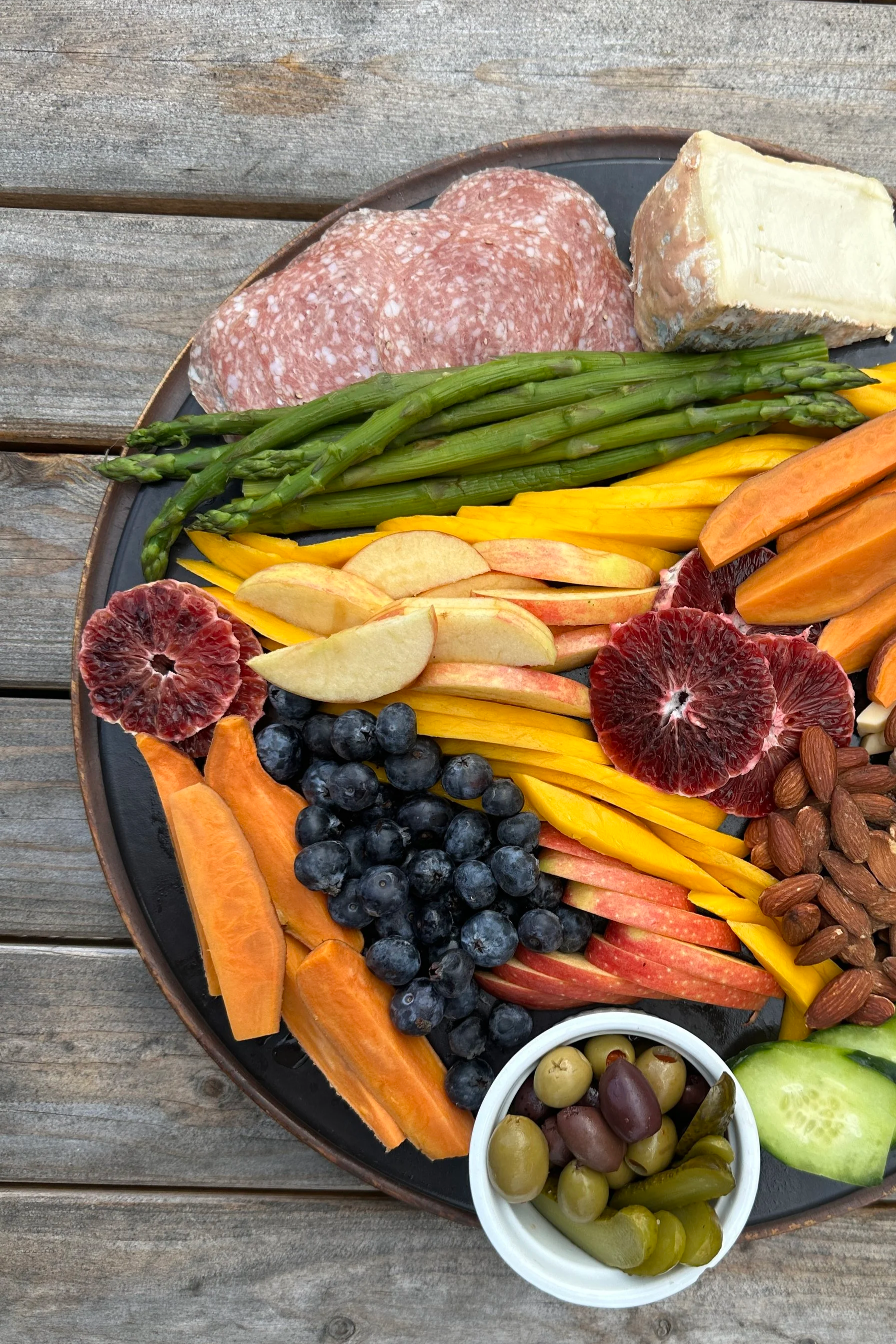 A round platter with a variety of sliced fruits, cheese, meats, and nuts on a wooden surface. Contains cantaloupe, blueberries, apples, grapefruit, grapes, almonds, and sliced salami, cheese, and vegetables.
