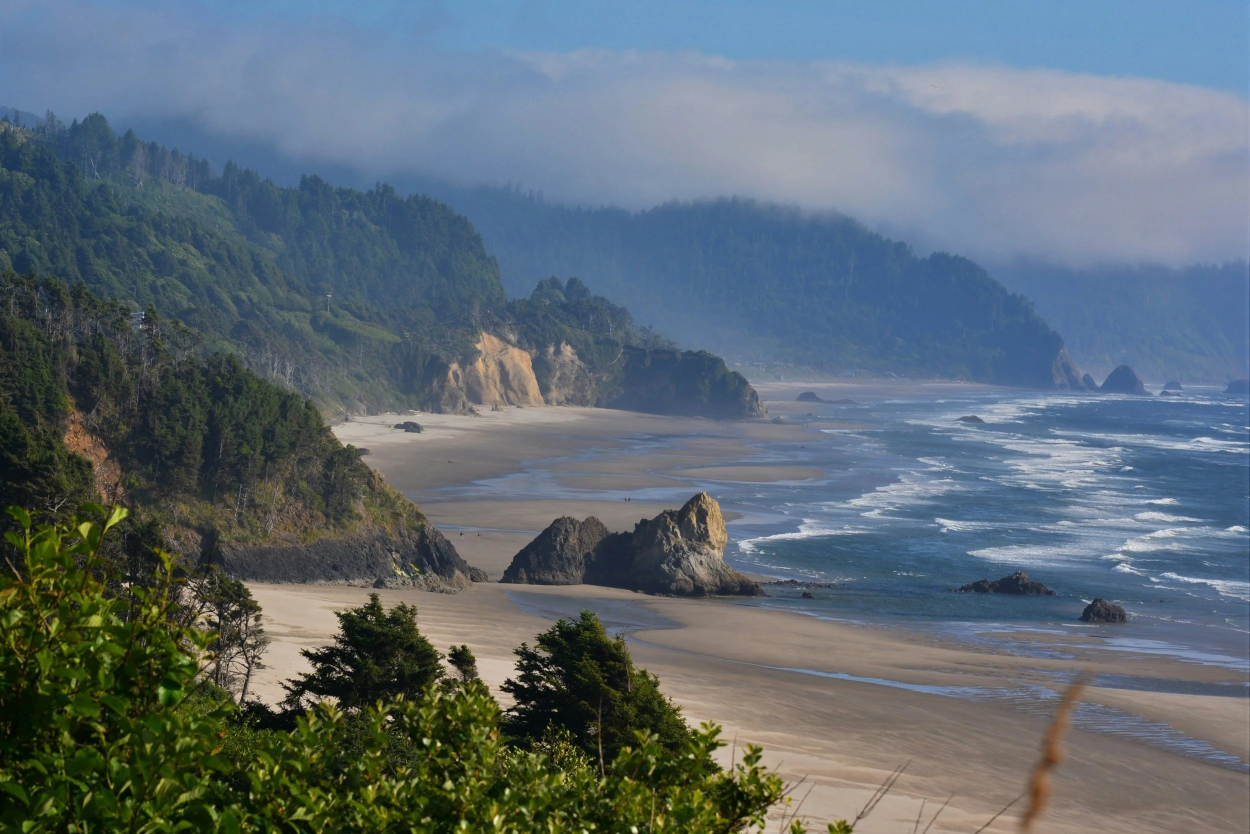 Coastal scene with sandy beach, rocky outcroppings, dense green forest on cliffs, misty mountains in the background, and the ocean with gentle waves.