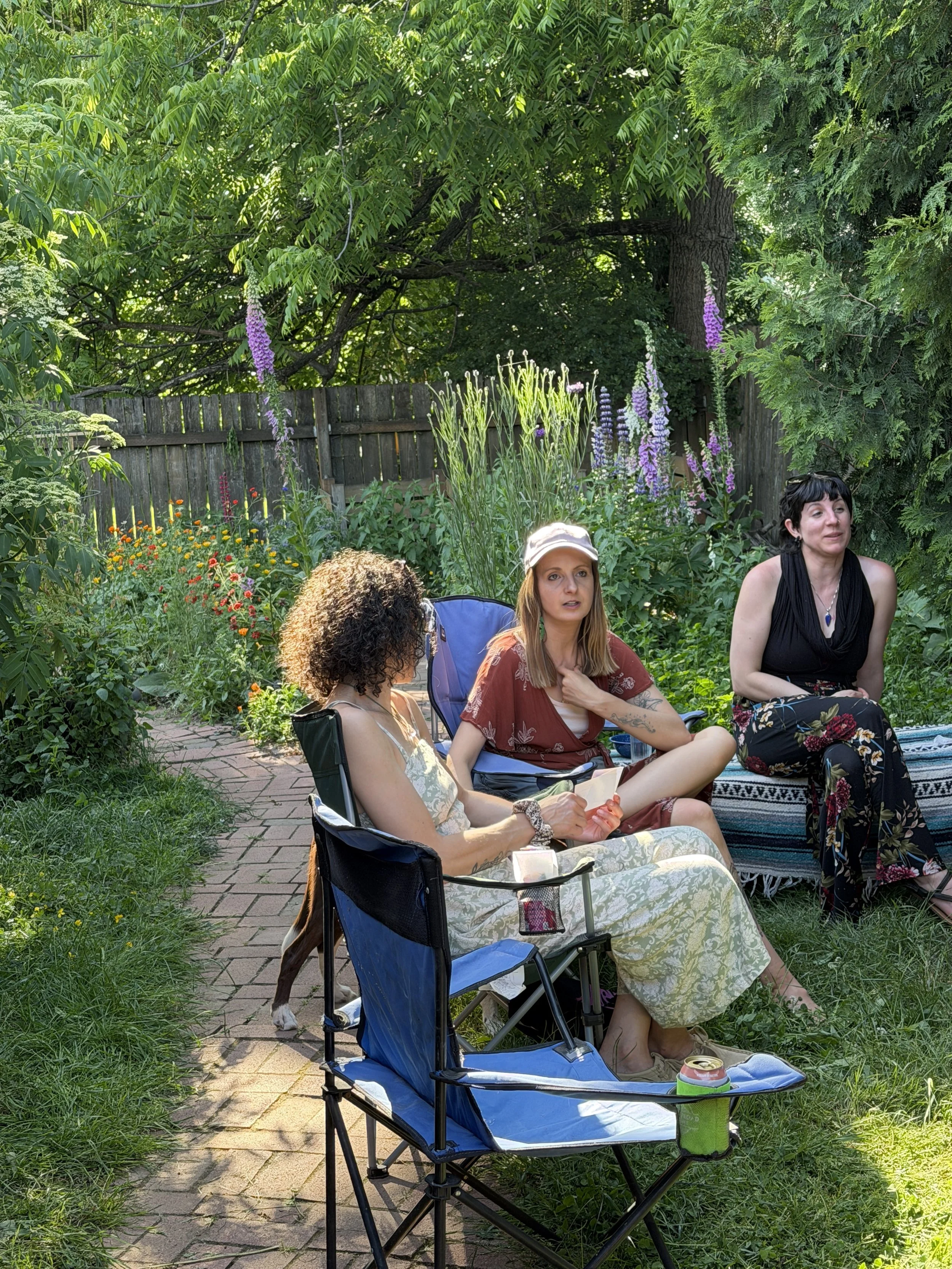 Three women sitting in a lush garden with a wooden fence and flowers, one holding a paper, a dog next to one woman, and a folding chair with a drink container in the foreground.