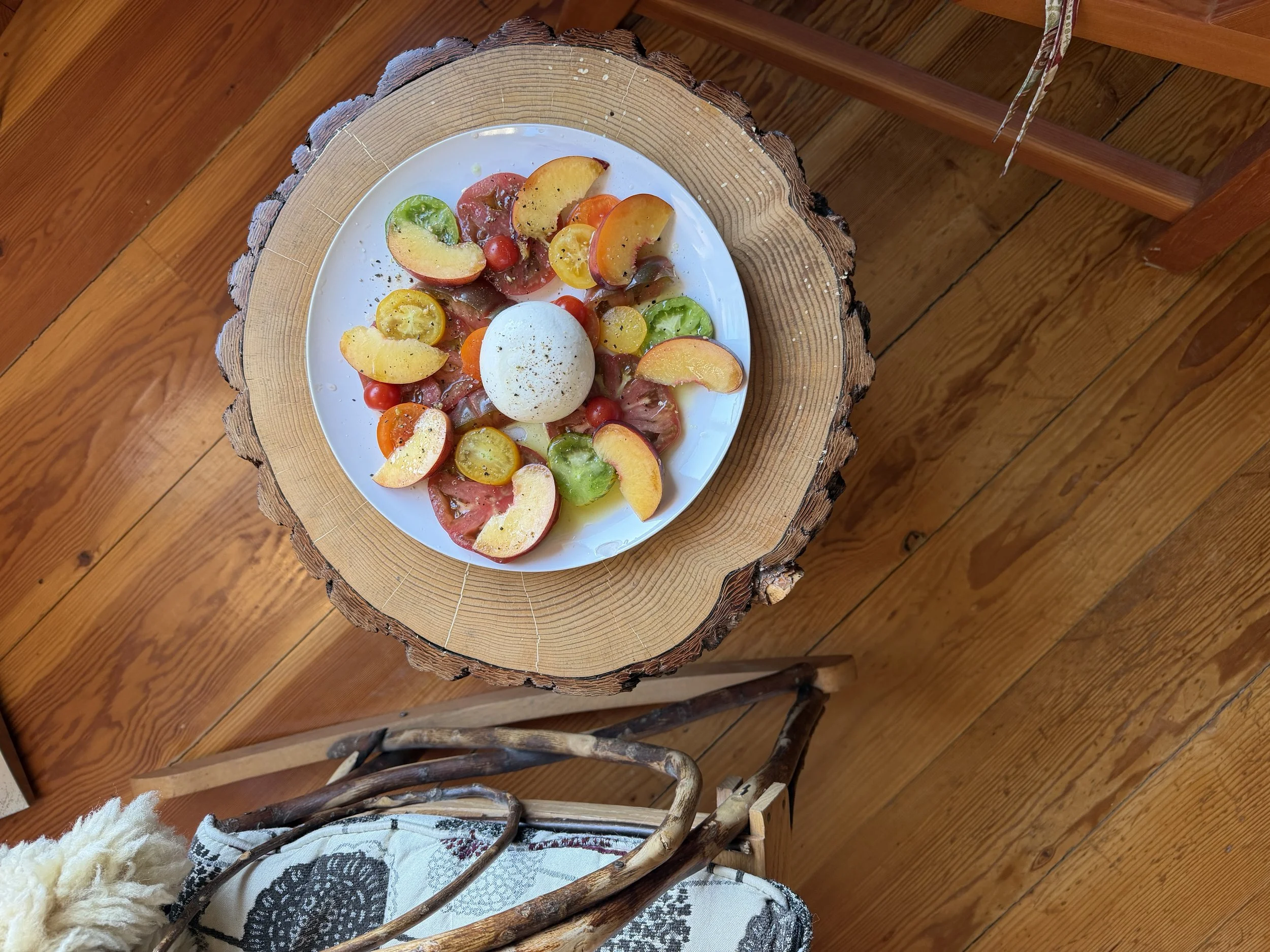 Close-up of a plate with assorted sliced fruits including peaches, grapes, cherry tomatoes, and a boiled egg, placed on a tree trunk coffee table with a wooden floor in the background.
