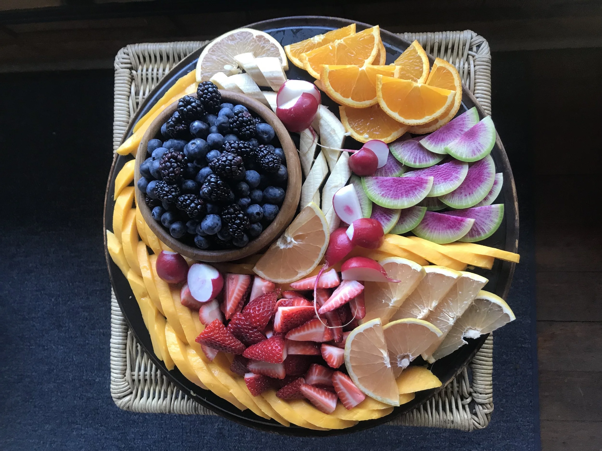 Plate of sliced fresh fruits including oranges, strawberries, watermelon, blueberries, blackberries, radishes, radish flowers, and lemon slices.