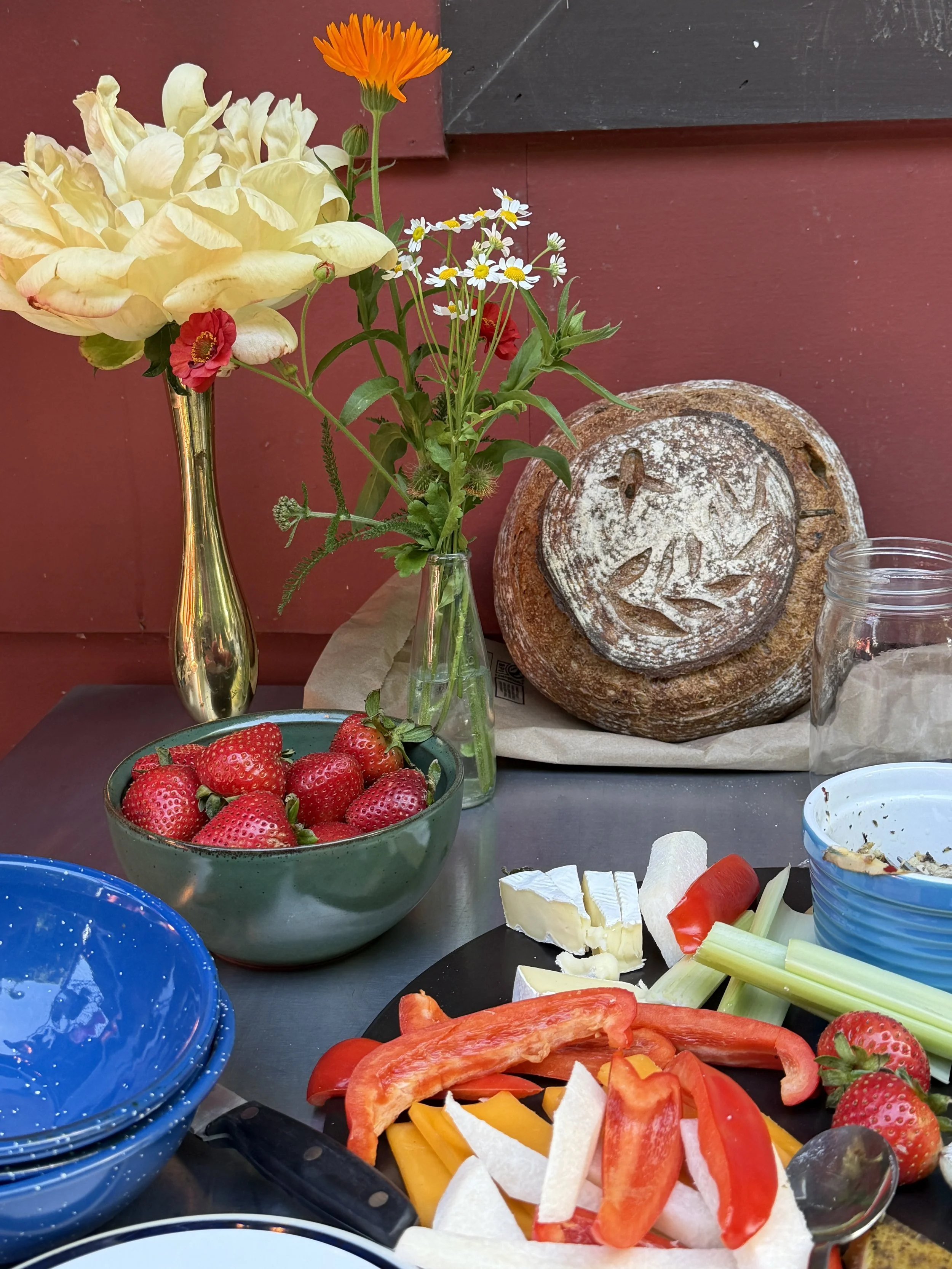 A table with a bowl of strawberries, sliced vegetables including bell peppers, celery, and melons, a loaf of bread, a cheese wedge, and a jar of seasoning, with flowers in a vase in the background.