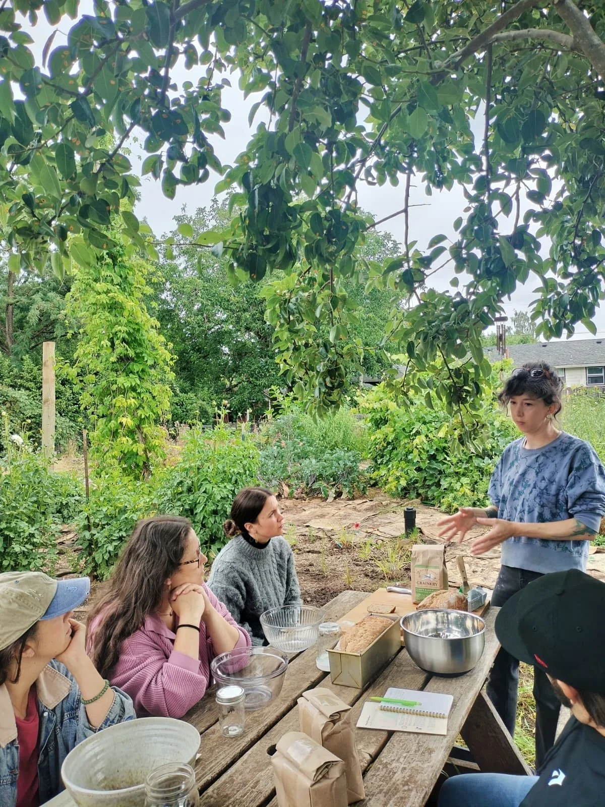 A group of people sitting at a wooden table outdoors in a garden, listening to a woman explain something. The table has bowls, jars, and paper bags on it. The background features lush green plants and trees.