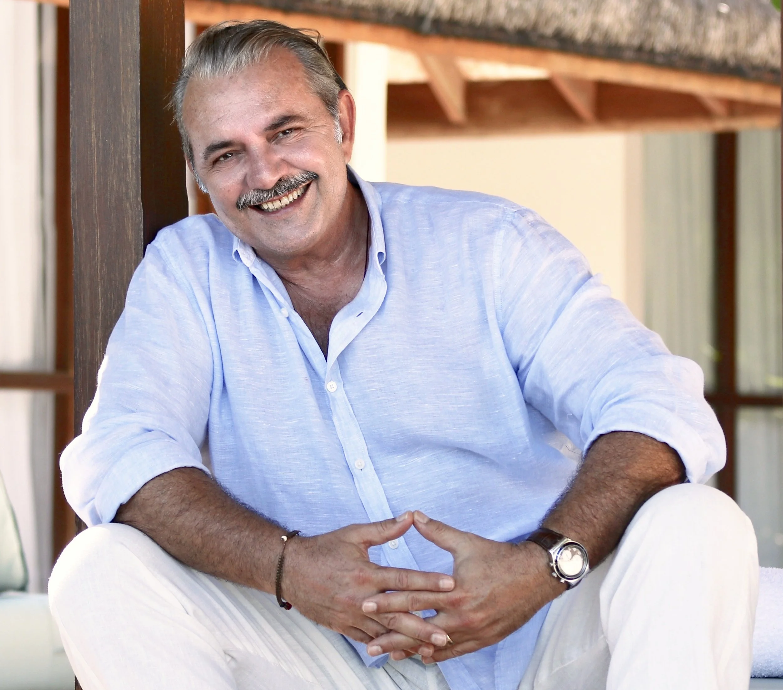 Smiling middle-aged man with gray hair and moustache, wearing a light blue shirt, sitting outdoors by wooden structure.